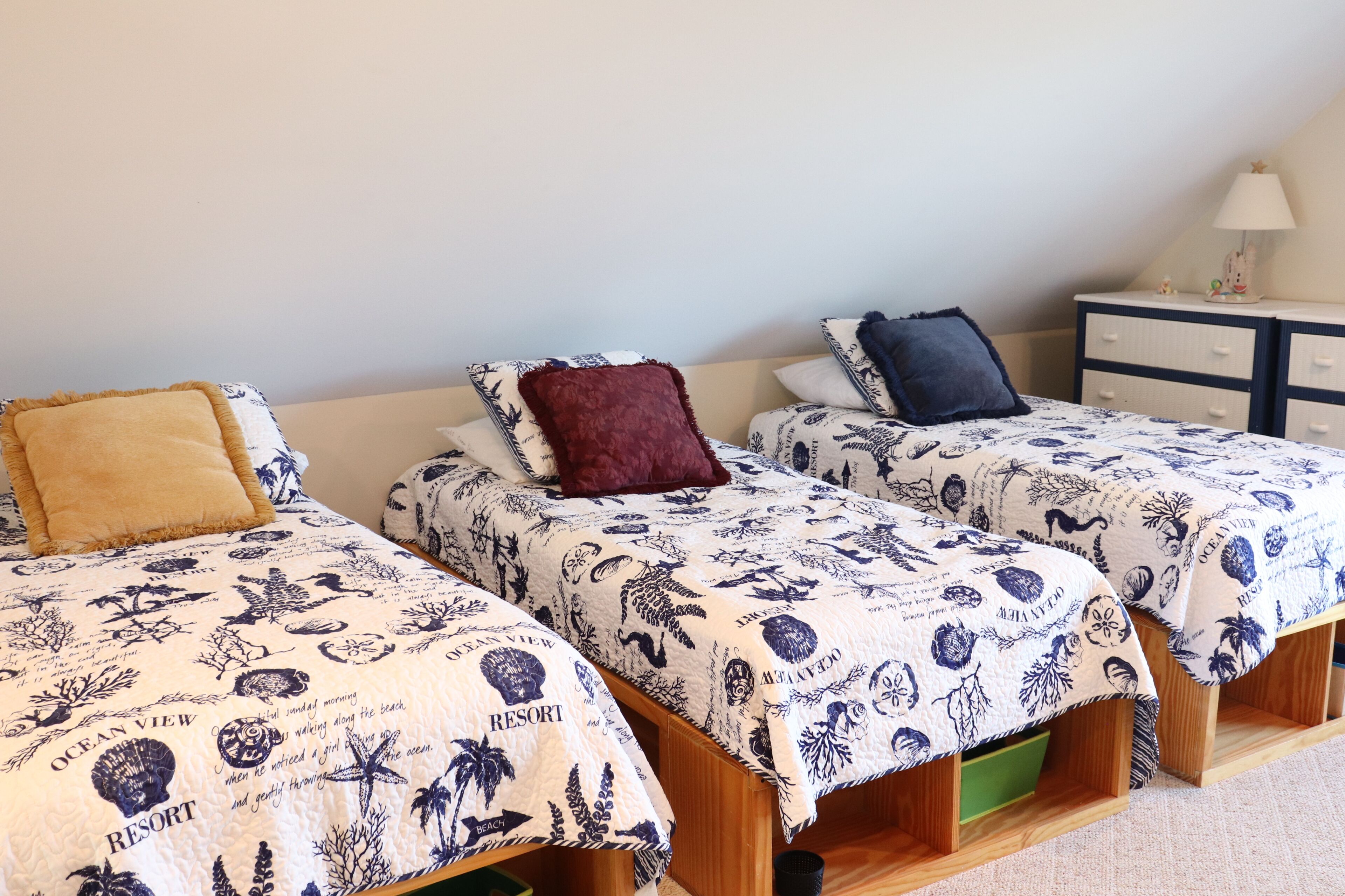 Trois lits simples avec la même literie bleue et blanche à motifs, une petite table de chevet avec une lampe et une commode avec des tiroirs.
