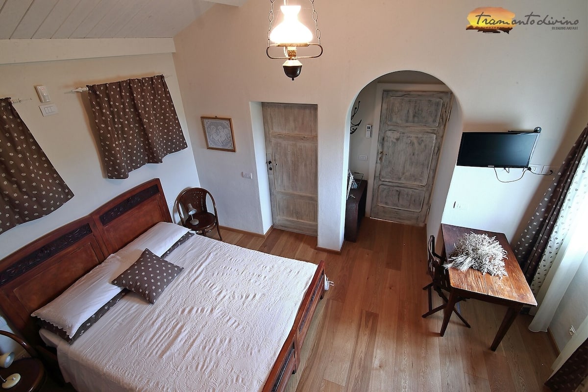 A bedroom with a wooden bed, a desk with a chair, a television, and a door leading to another room.
