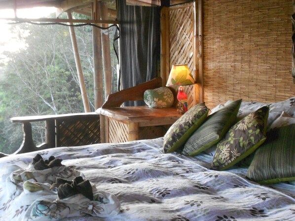 Una habitación con cama, ventana con vista a árboles, lámpara y pared con tejido.