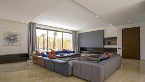 A spacious living area with a large sectional sofa, a fireplace, and a view of palm trees through large windows. - Villa Amara Oliver Palm Resort (Marrakech)