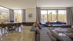 A spacious living room with a large sofa, a dining table with chairs, and a view of the outdoors through large windows. - Villa Amara Oliver Palm Resort (Marrakech)