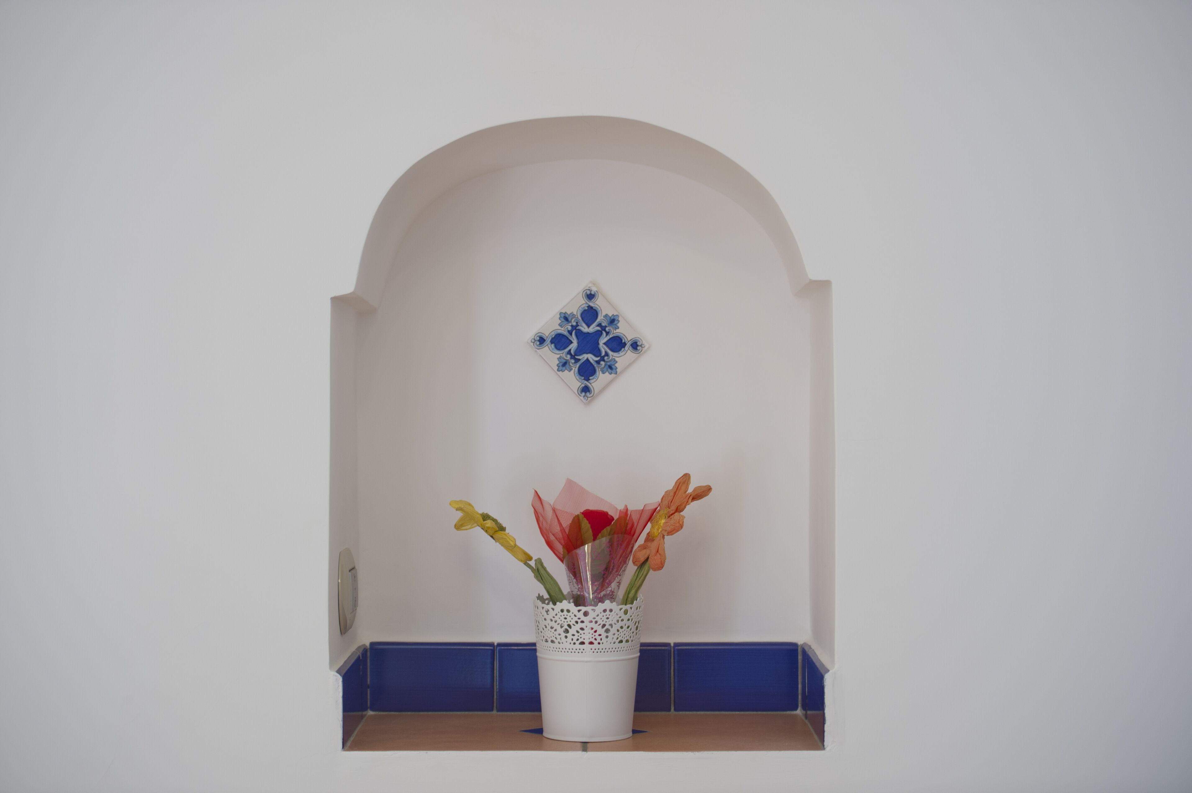 A white wall with a blue-tiled baseboard, a white decorative tile above a white vase with flowers, and a small shelf below.