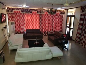 A living room with a white sofa, a brown sofa, and a black office chair. There is a ceiling fan and a chandelier. - Nass Lodge (Sunyani)