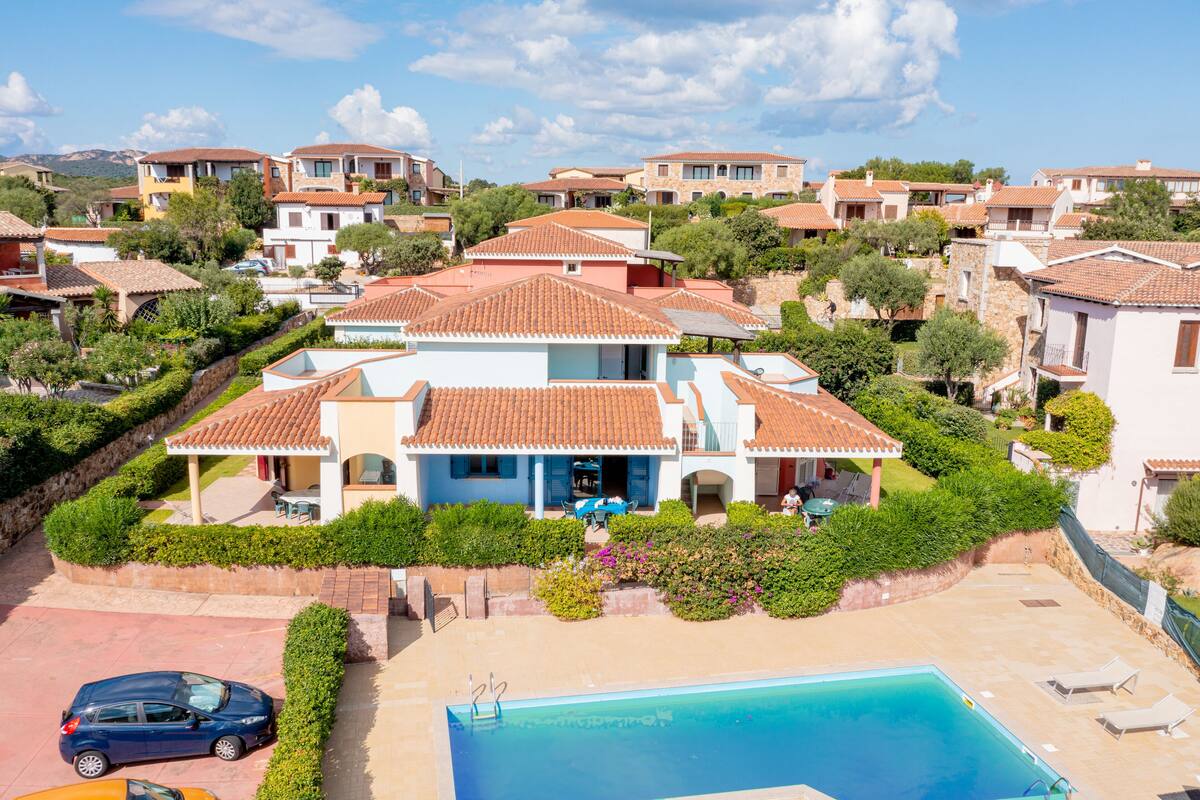 A large villa with a swimming pool, surrounded by lush greenery and other residential buildings.