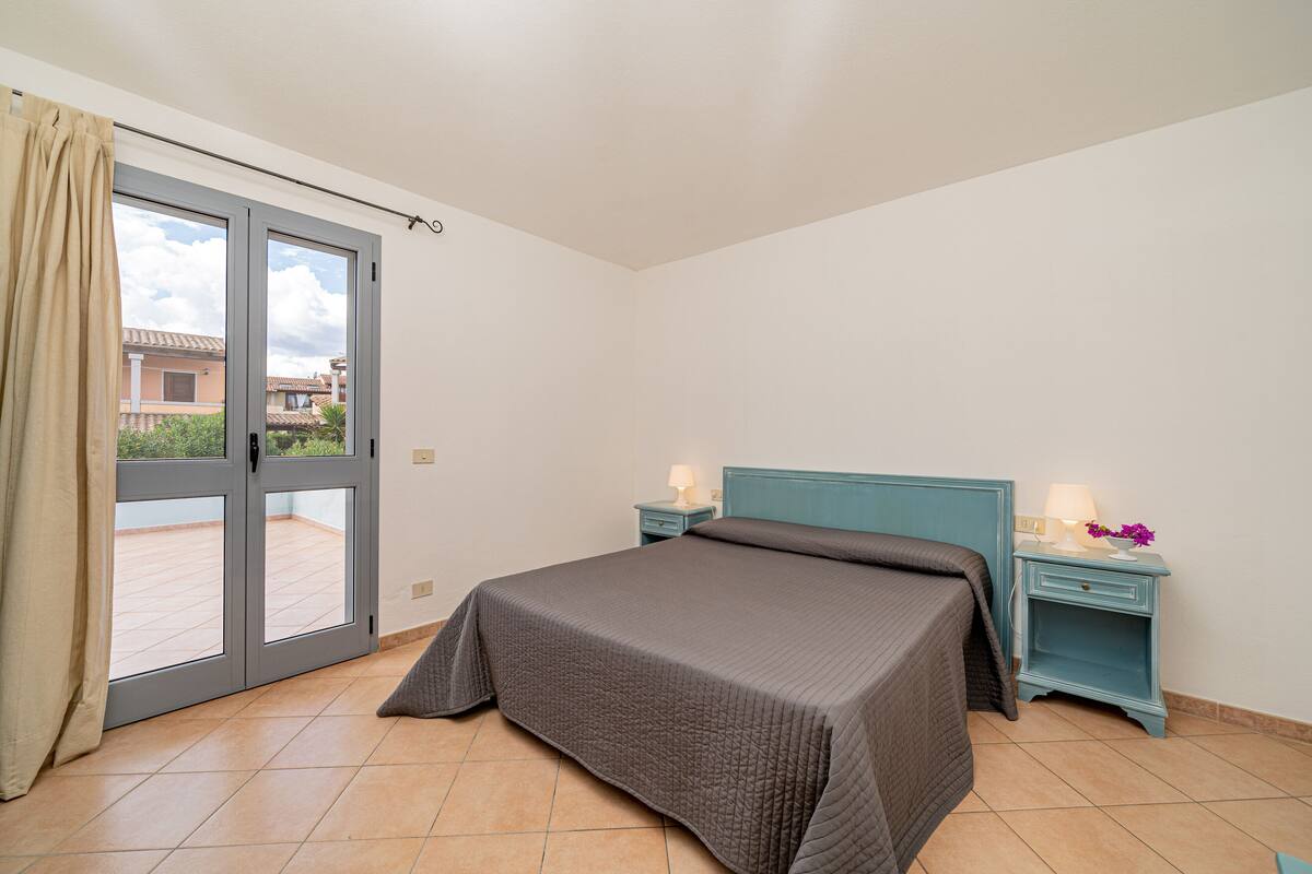 A bedroom with a bed, a nightstand, a window with curtains, and a view of the outside.
