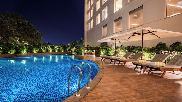 A swimming pool with lounge chairs and umbrellas, surrounded by greenery and a building with large windows. - Lemon Tree Premier Mumbai Intl. Airport (Mumbai)