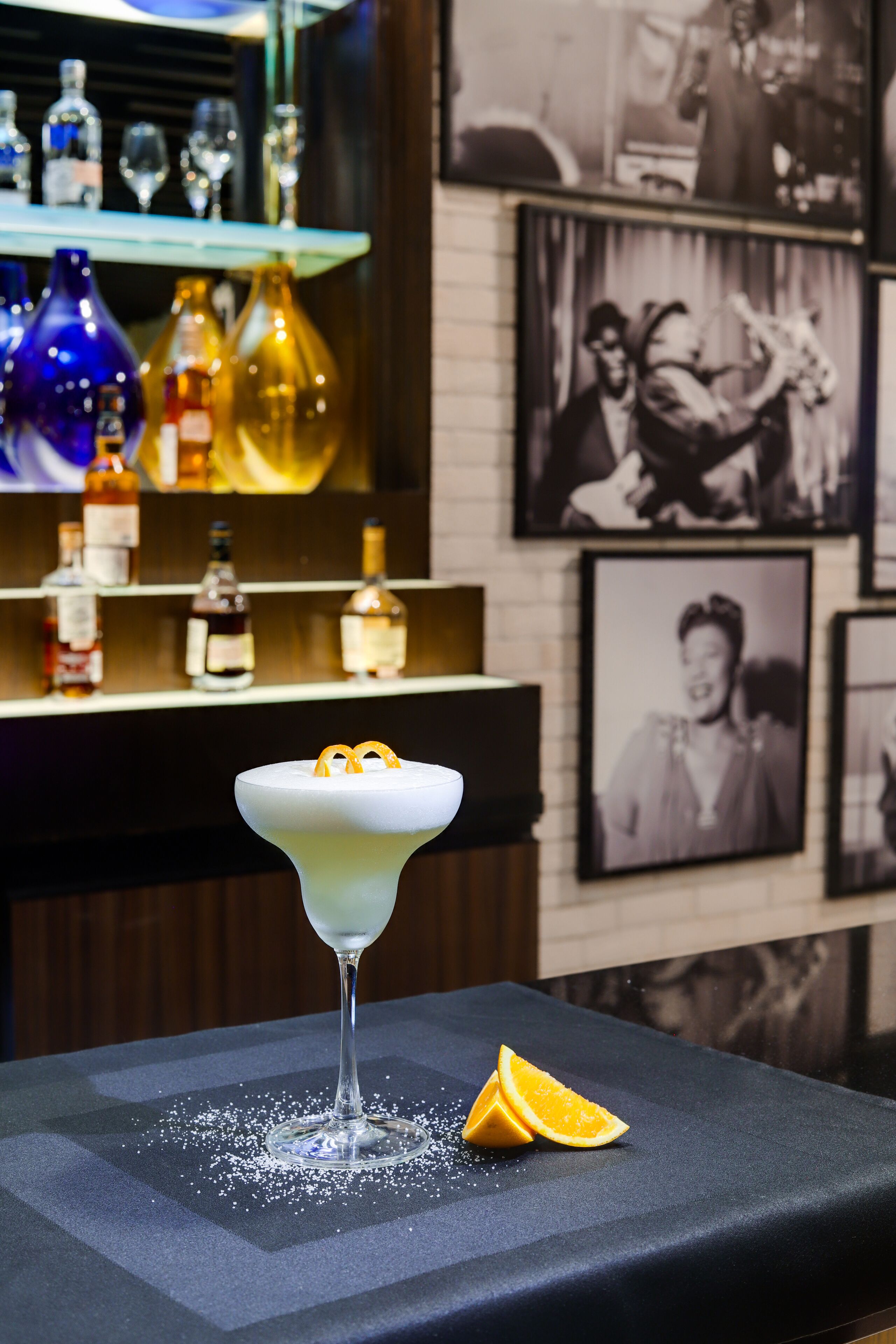 A bar area with a cocktail on a table, bottles in the background, and framed pictures on the wall.
