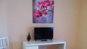 Une pièce avec un bureau blanc, un téléviseur, une petite plante en pot, une chaise blanche et un lit.