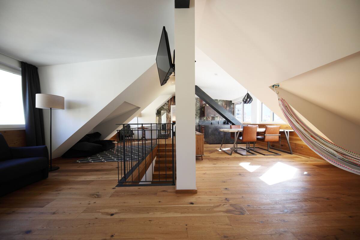 A spacious loft with a wooden floor, a staircase, a dining area, and a hammock.