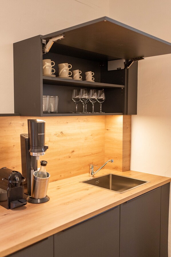 A modern kitchen with a built-in shelf holding cups and glasses, a coffee machine, and a sink. - Residence Rösch (Merano)