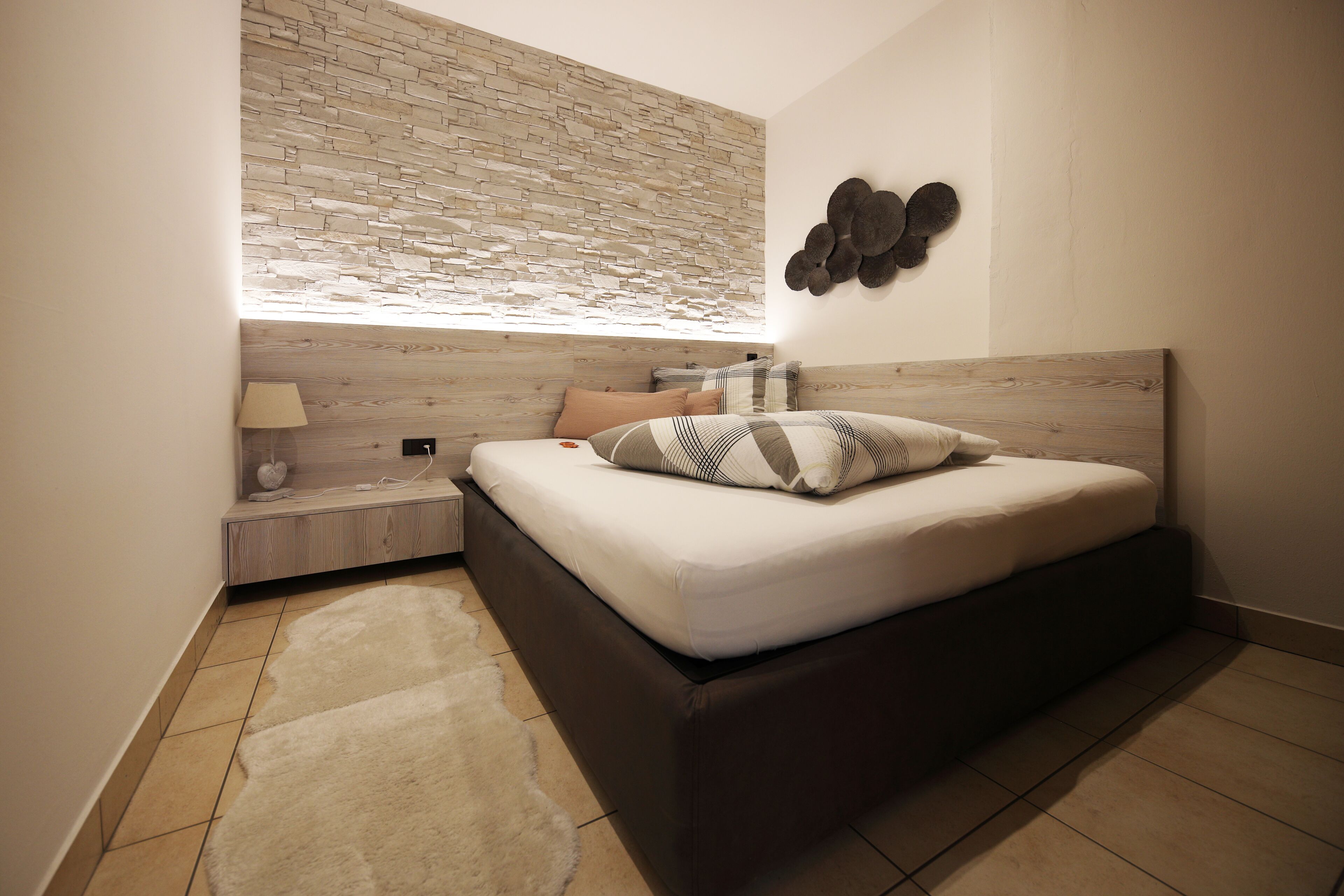 Un dormitori amb un llit, una tauleta de nit, una làmpada, un rellotge de paret, un element decoratiu a la paret i una finestra amb vista a l'exterior.
