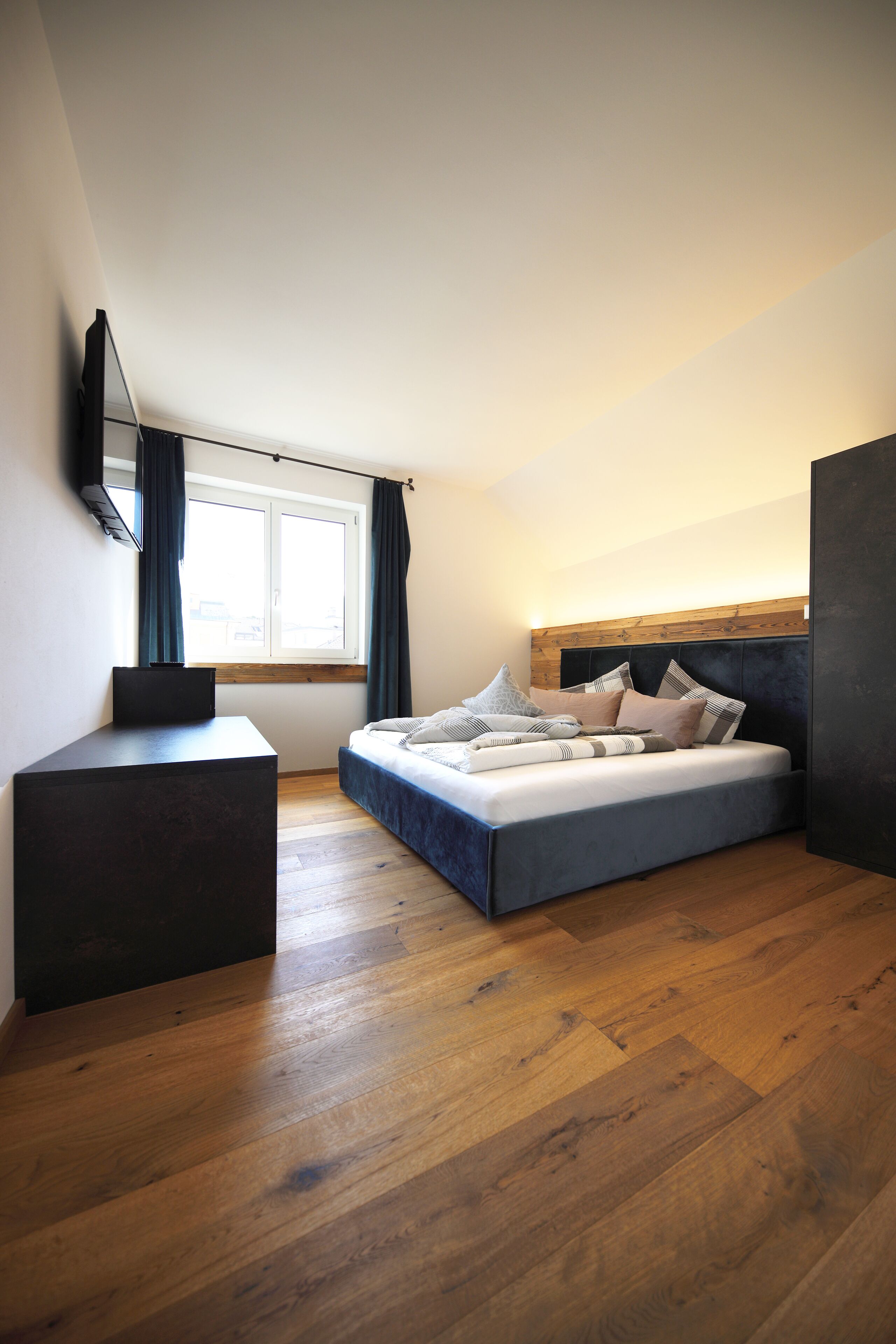 A bedroom with a bed, a desk, a window with curtains, and a wooden floor.