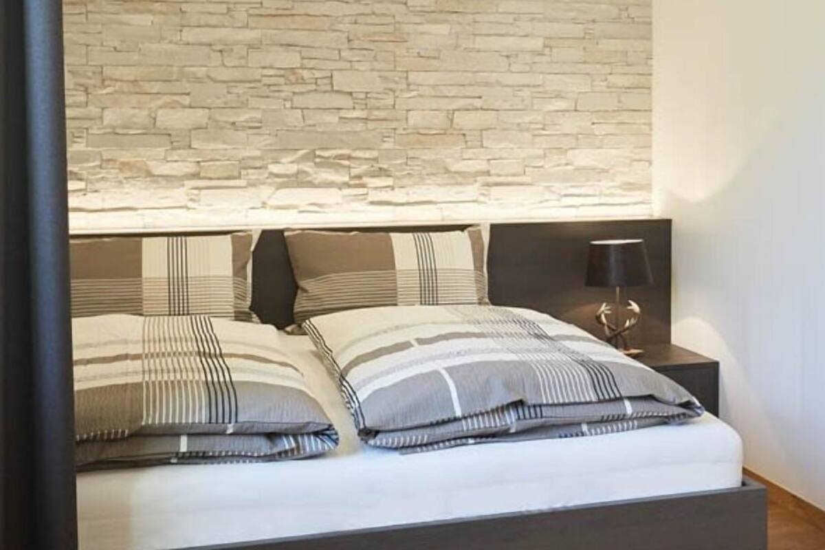 A bed with striped bedding, a stone wall behind it, and a nightstand with a lamp.