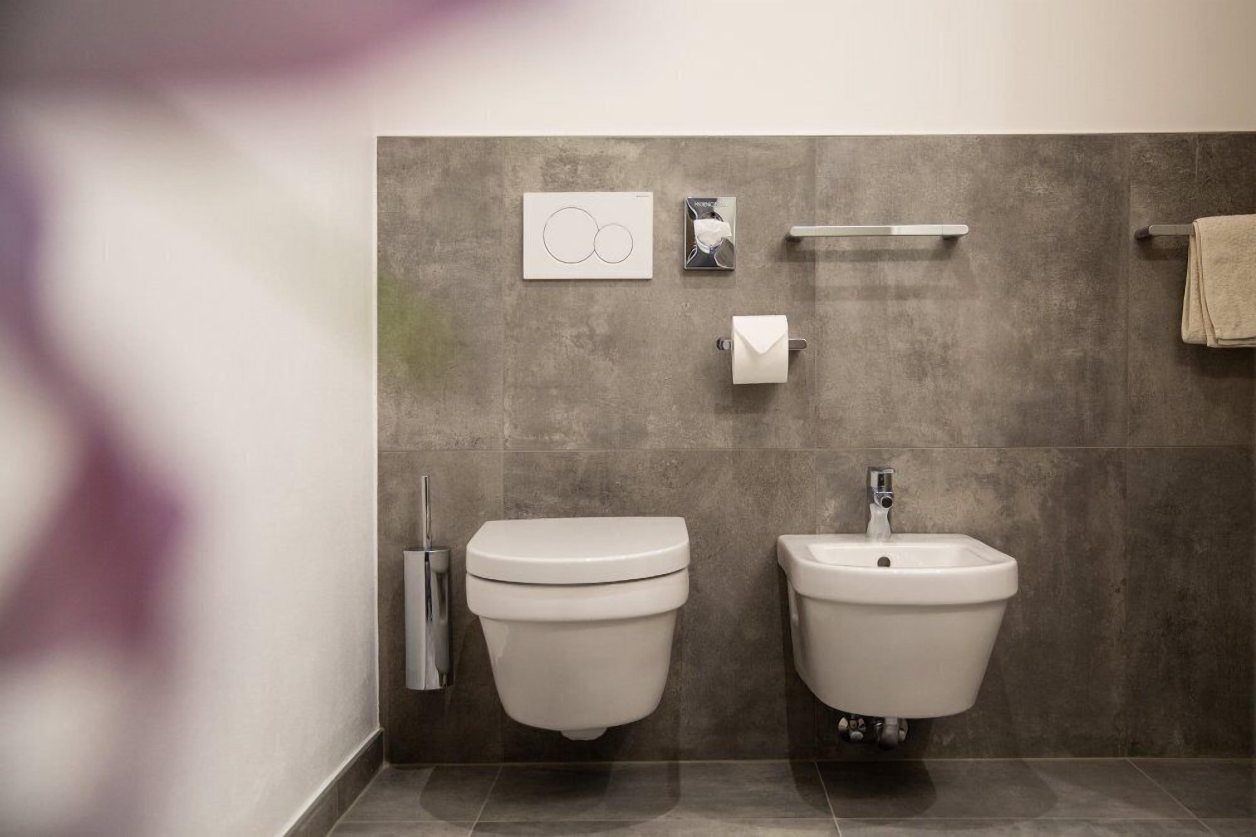 Et moderne badeværelse med to toiletter og en bidet, et håndklædestativ samt en sæbedispenser på væggen.