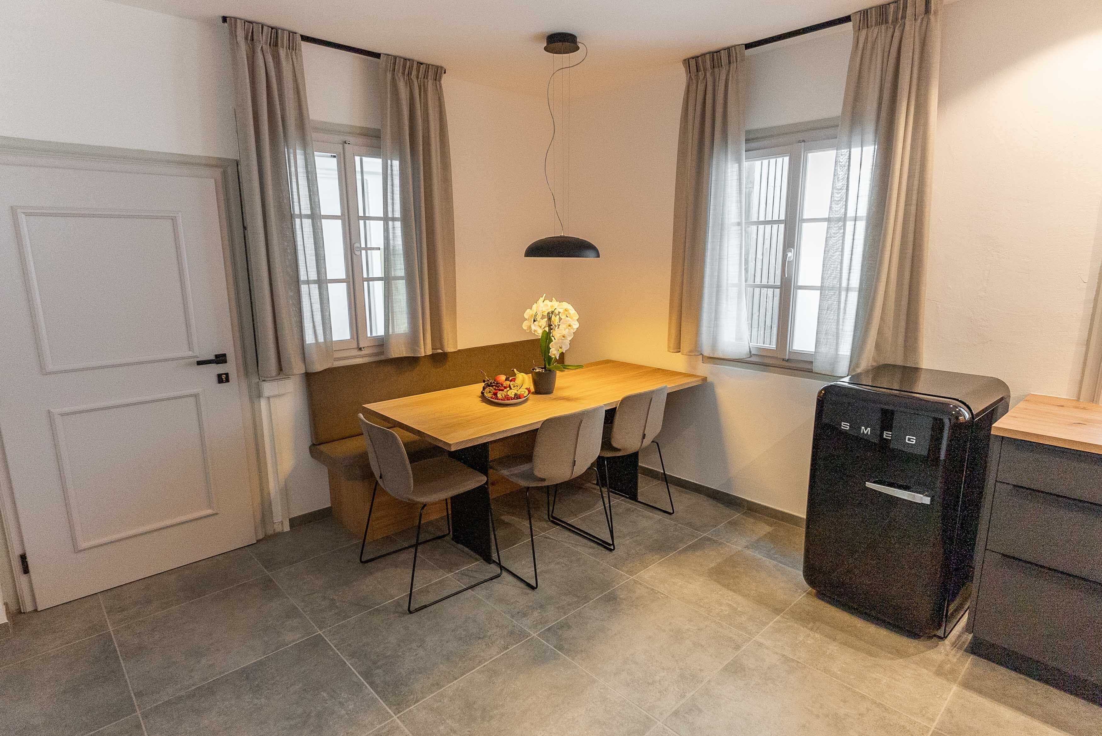 A compact kitchen with a wooden table, chairs, a small refrigerator, and a vase with flowers.