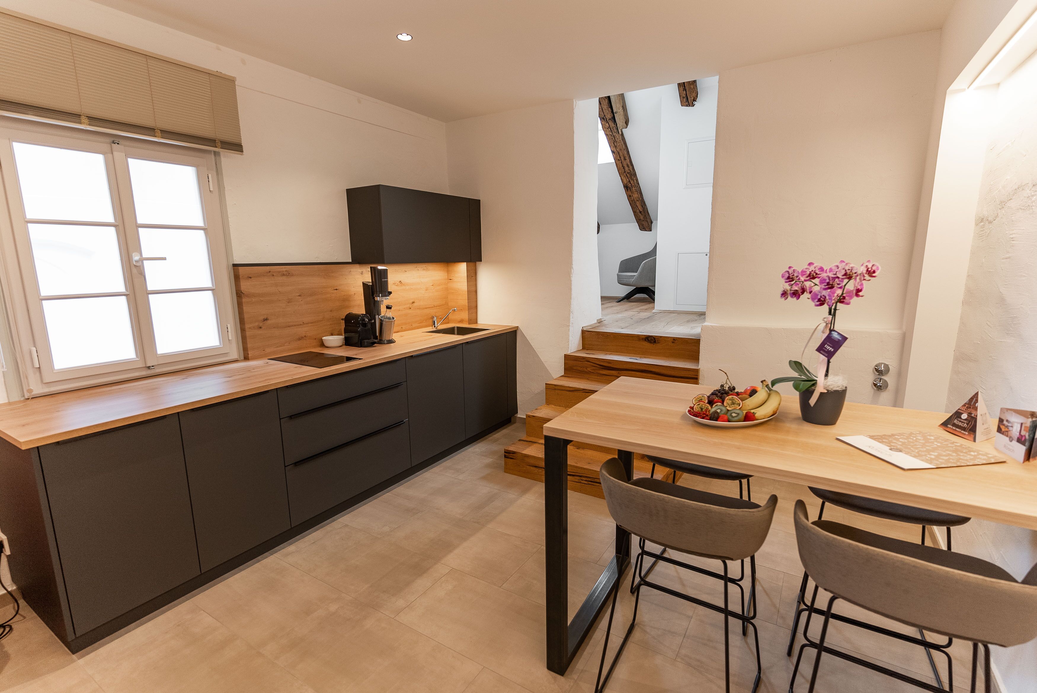 A modern kitchen with dark cabinets, a wooden countertop, and a built-in stove. There is a dining area with a wooden table and bar stools.