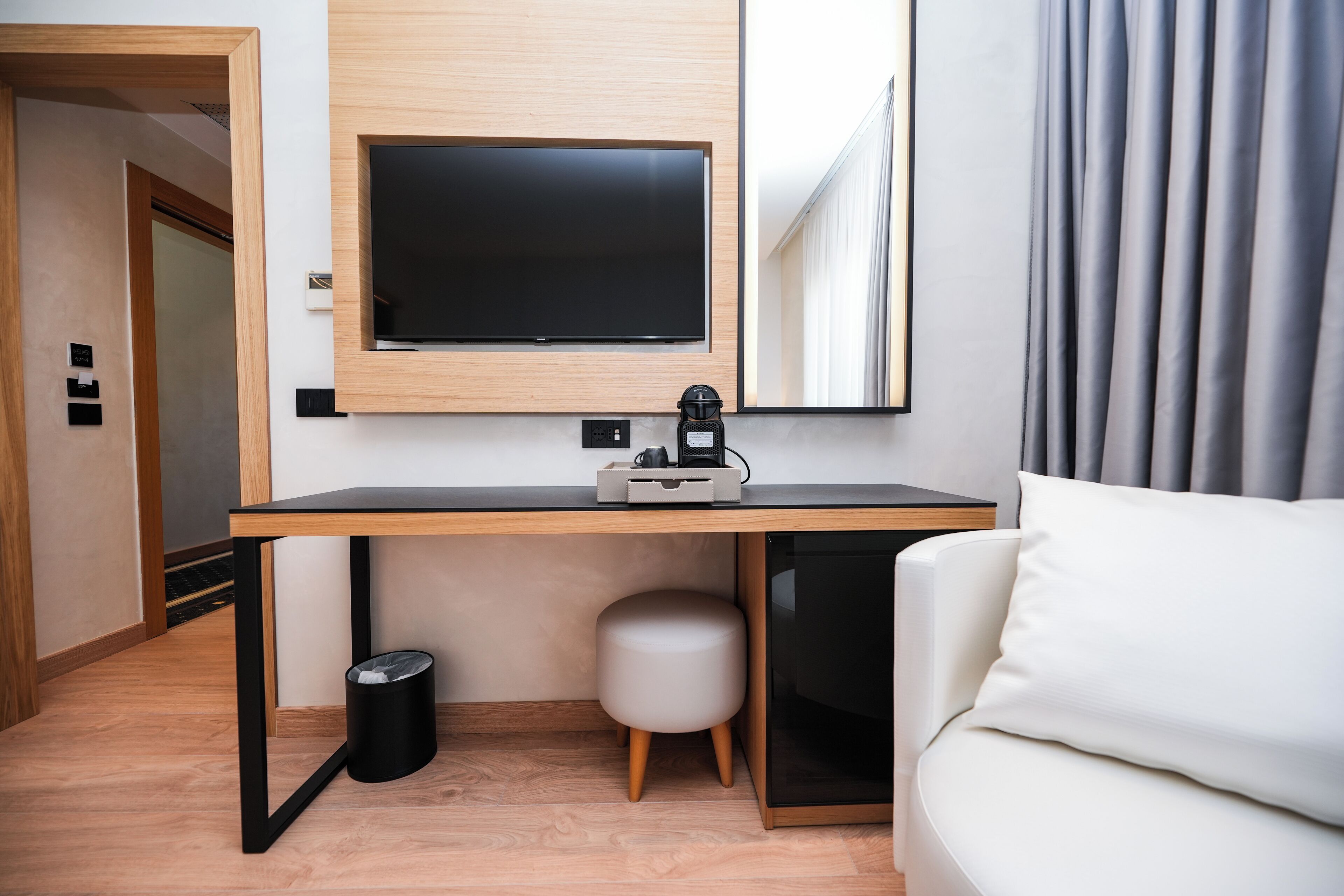 Ein modernes Hotelzimmer mit Schreibtisch aus Holz, einem Flachbildfernseher, einem kleinen Hocker und einem weißen Sofa.