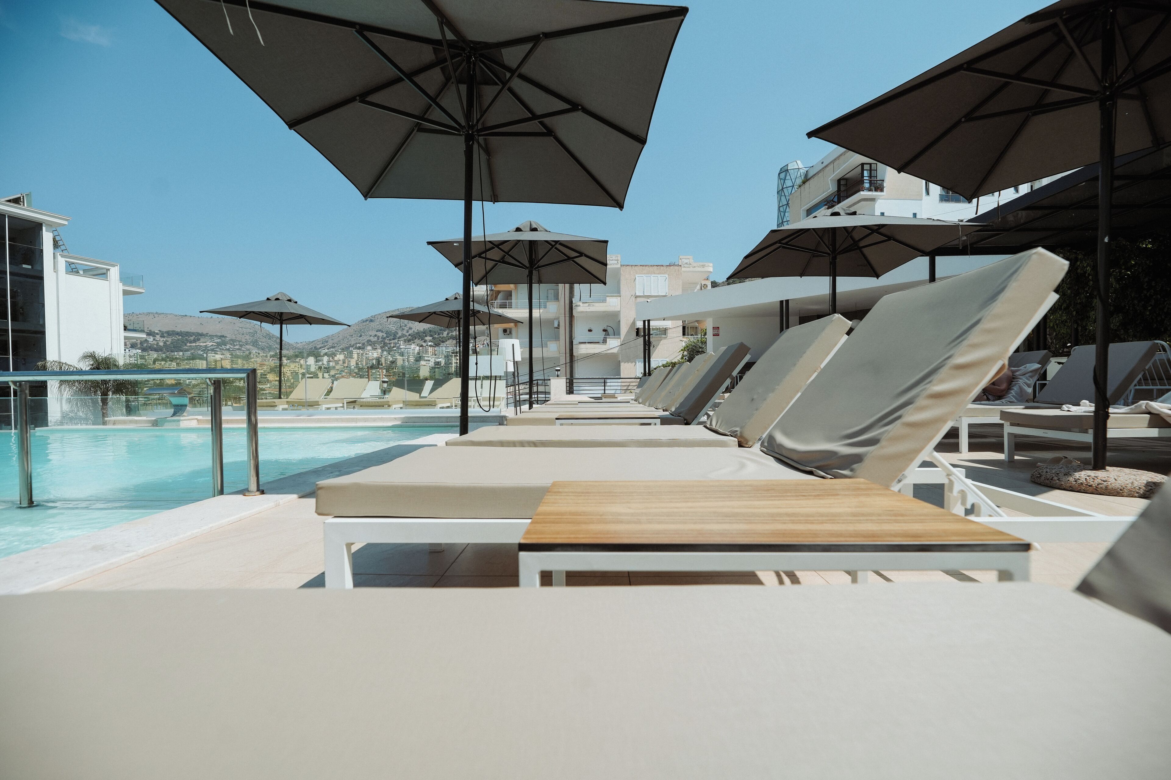 A poolside area with lounge chairs and umbrellas, a clear view of the pool, and modern buildings in the background.