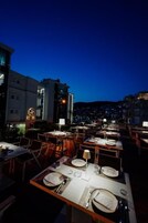 Un área de comedor al aire libre con mesas dispuestas para dos, con vistas a un paisaje urbano al atardecer.