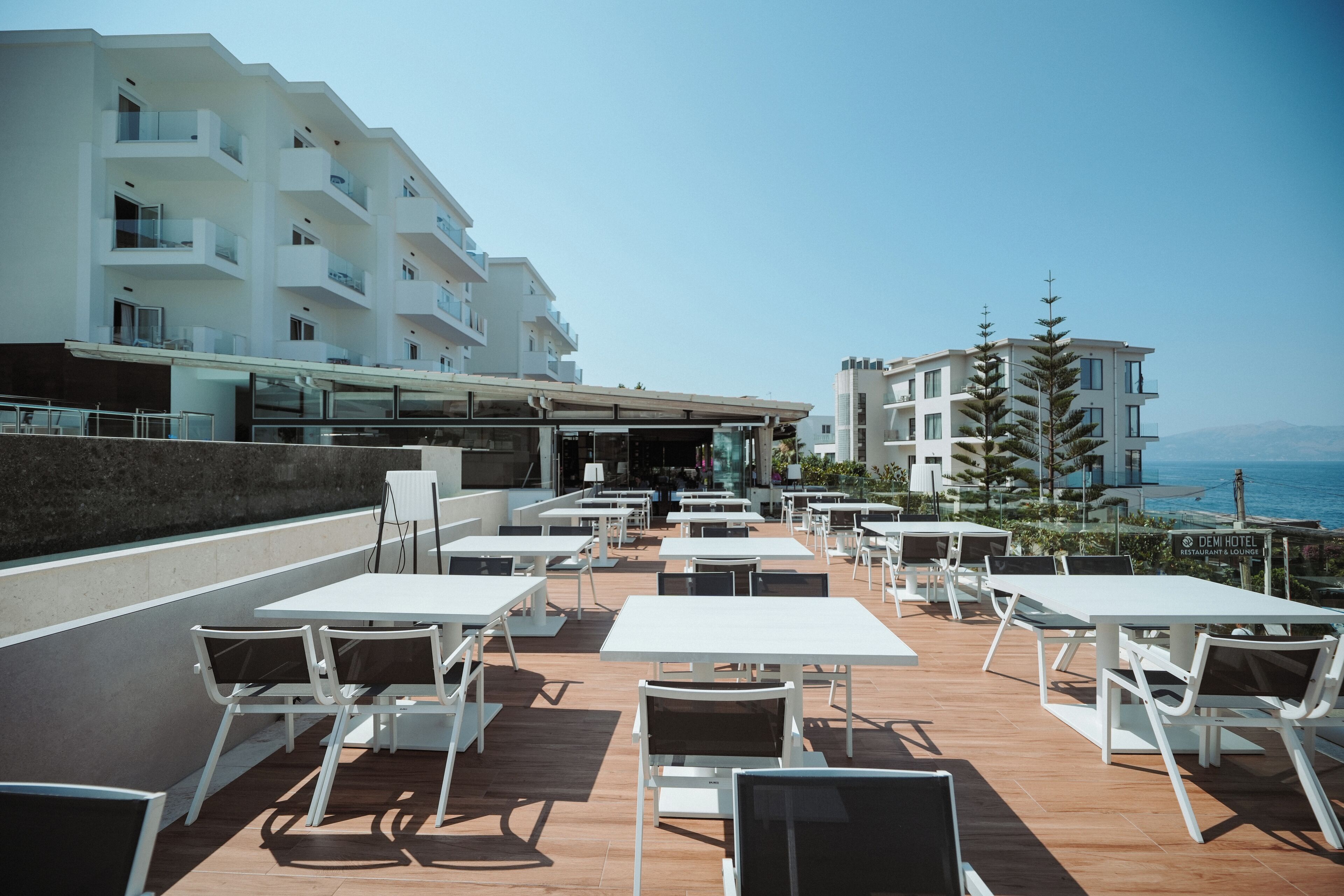An outdoor patio with white tables and chairs, overlooking a body of water and a building with balconies.