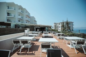 An outdoor patio with white tables and chairs, overlooking a body of water and a building with balconies.