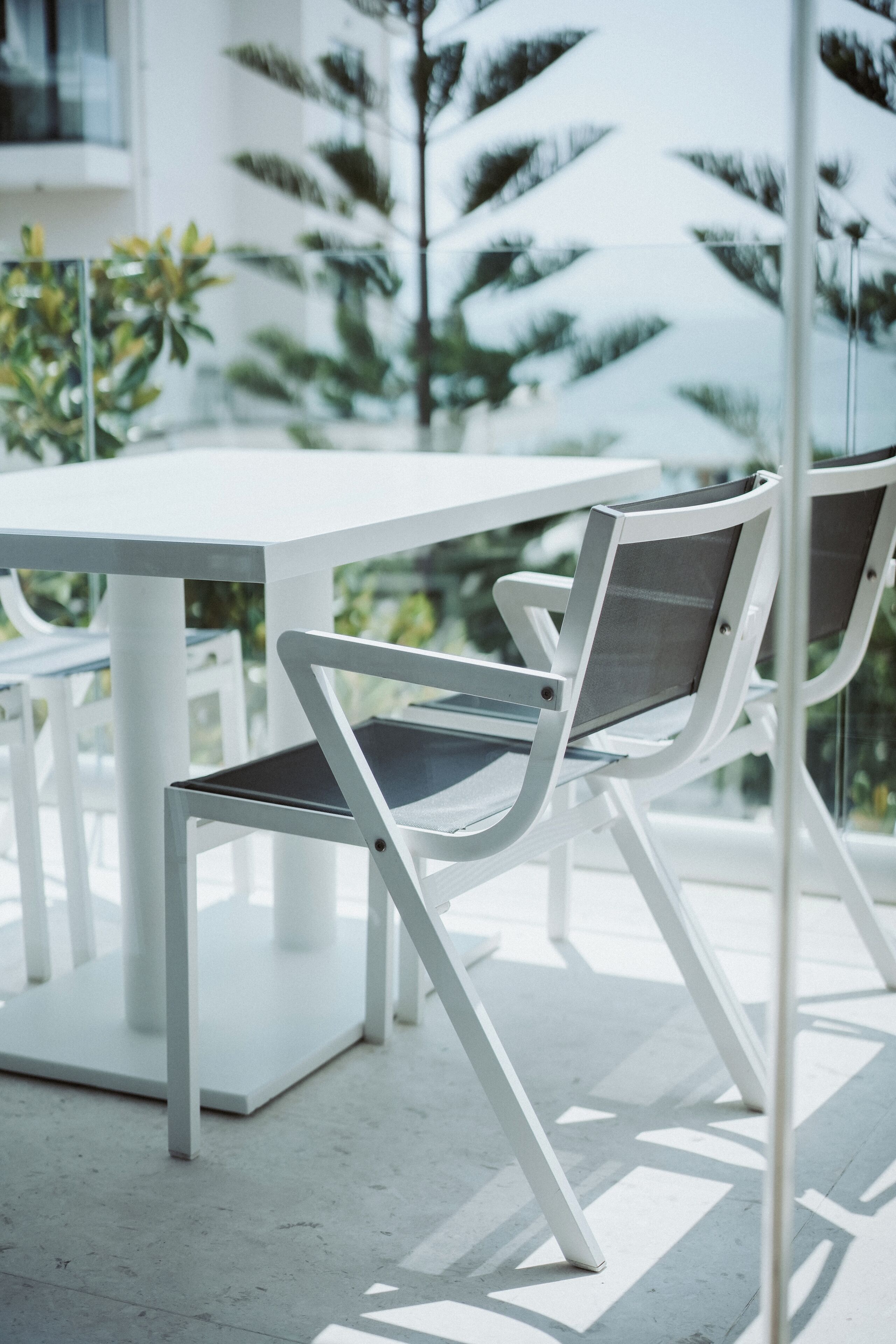 A modern outdoor seating area with a white table and chairs, surrounded by glass walls and greenery.