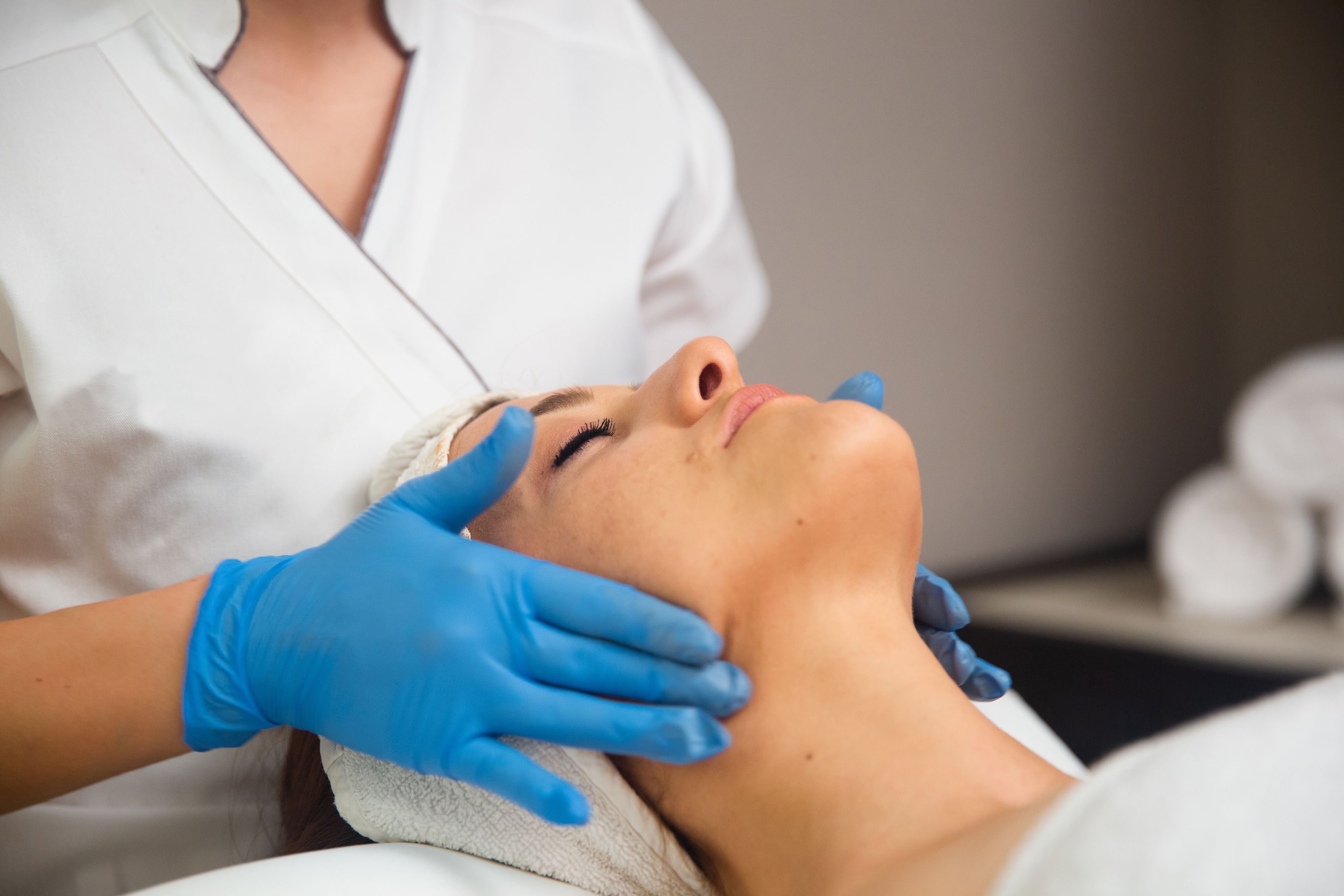 A person lying down with a towel on their head, receiving a facial treatment from a professional wearing blue gloves.