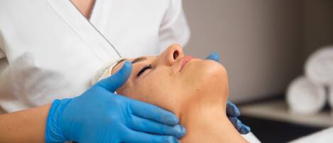 Una persona recostada con una toalla en la cabeza, recibiendo un tratamiento facial de un profesional que usa guantes azules.