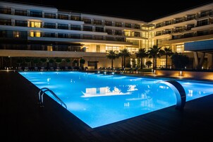 Un hotel moderno con una amplia piscina iluminada, varios balcones y palmeras.