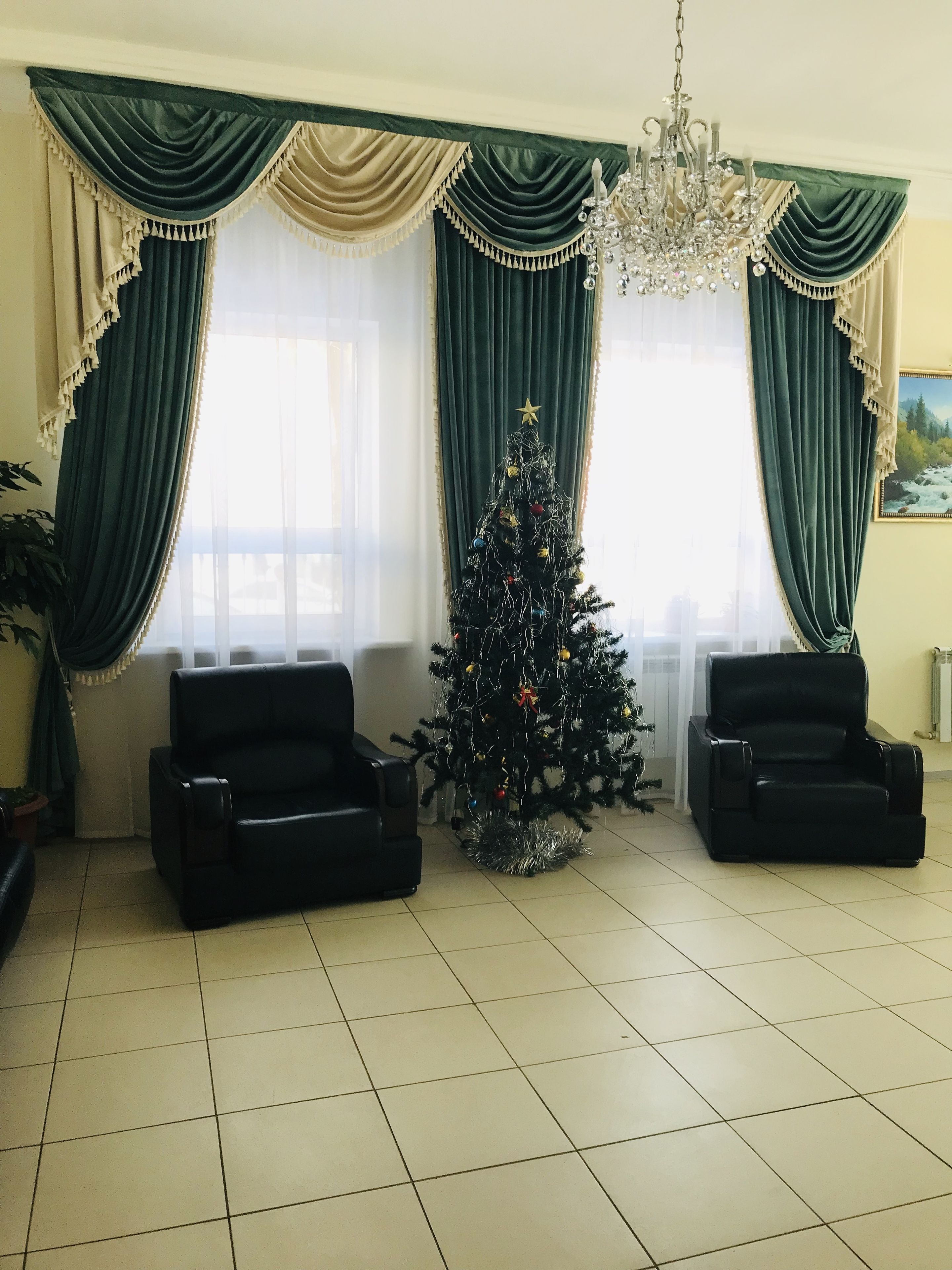 A living room with a Christmas tree, two black armchairs, a chandelier, and green curtains.