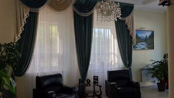 A living room with two black leather chairs, a small table, and a chandelier. There are curtains and a painting on the wall.