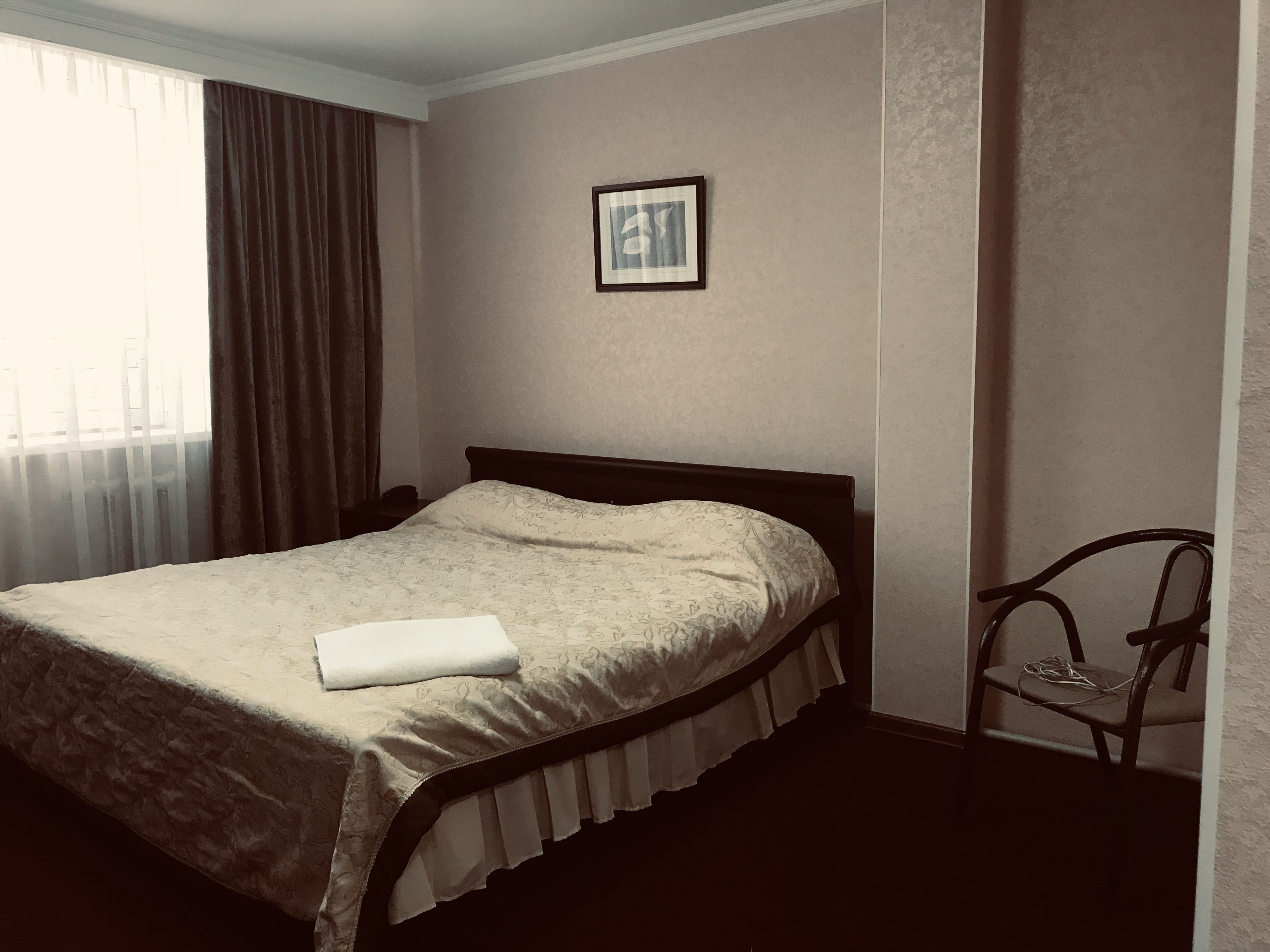 Una cama bien tendida con un edredón blanco y una toalla blanca doblada, un pequeño cuadro en la pared y una silla con un cable.
