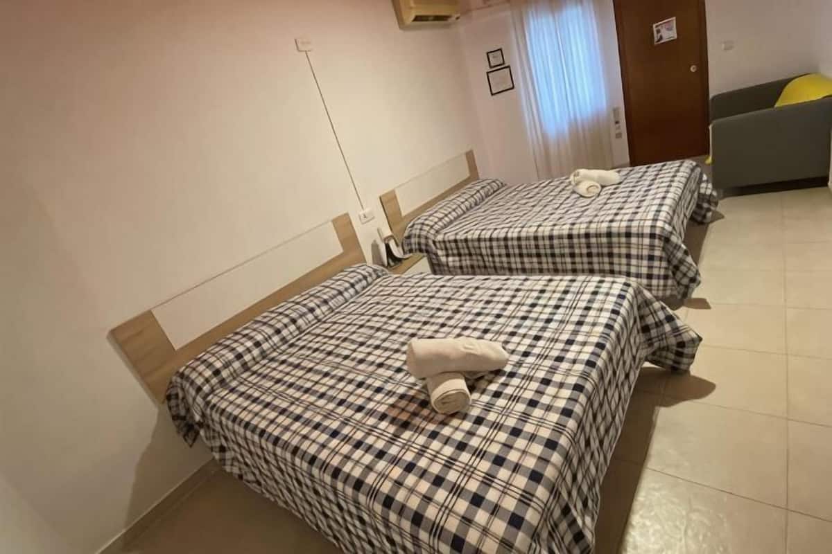 Kaksi sänkyä ruutukuosisilla vuodevaatteilla, yöpöytä, tuoli ja ovi hotellihuoneessa.