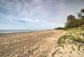 A sandy beach with footprints leading to the water, a distant pier, and trees on the right. - Geneva Cottage w/ Patio - Walk to Park, Lake Erie! (Geneva-on-the-Lake)