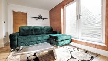 A modern living room with a green sectional sofa, a coffee table, and a large window with blinds.