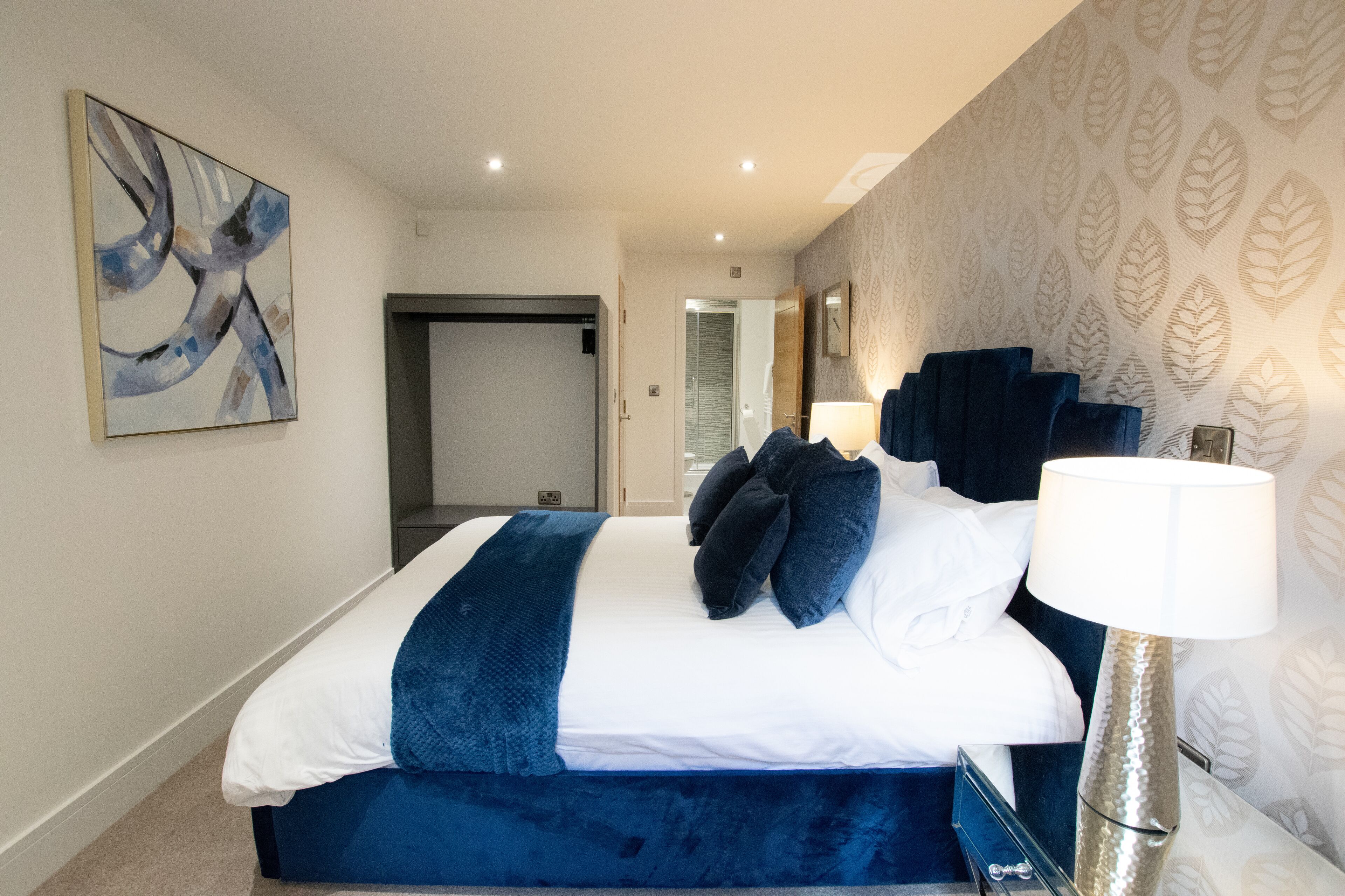 A modern hotel room with a large bed, a blue accent wall, a decorative lamp, and a leaf-patterned wallpaper.