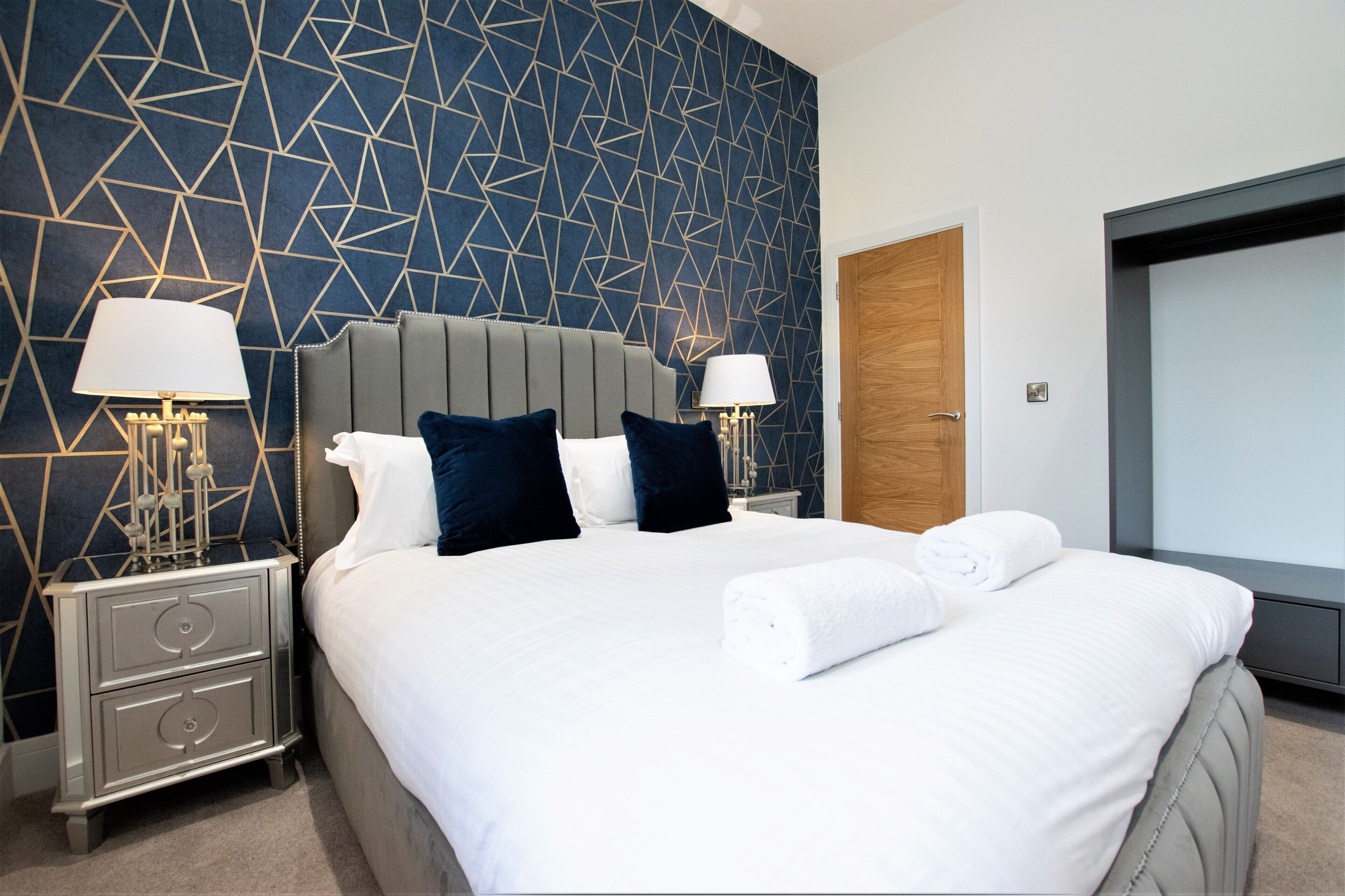 A hotel room with a large bed, two bedside tables with lamps, a wooden door, and a geometric-patterned wall.
