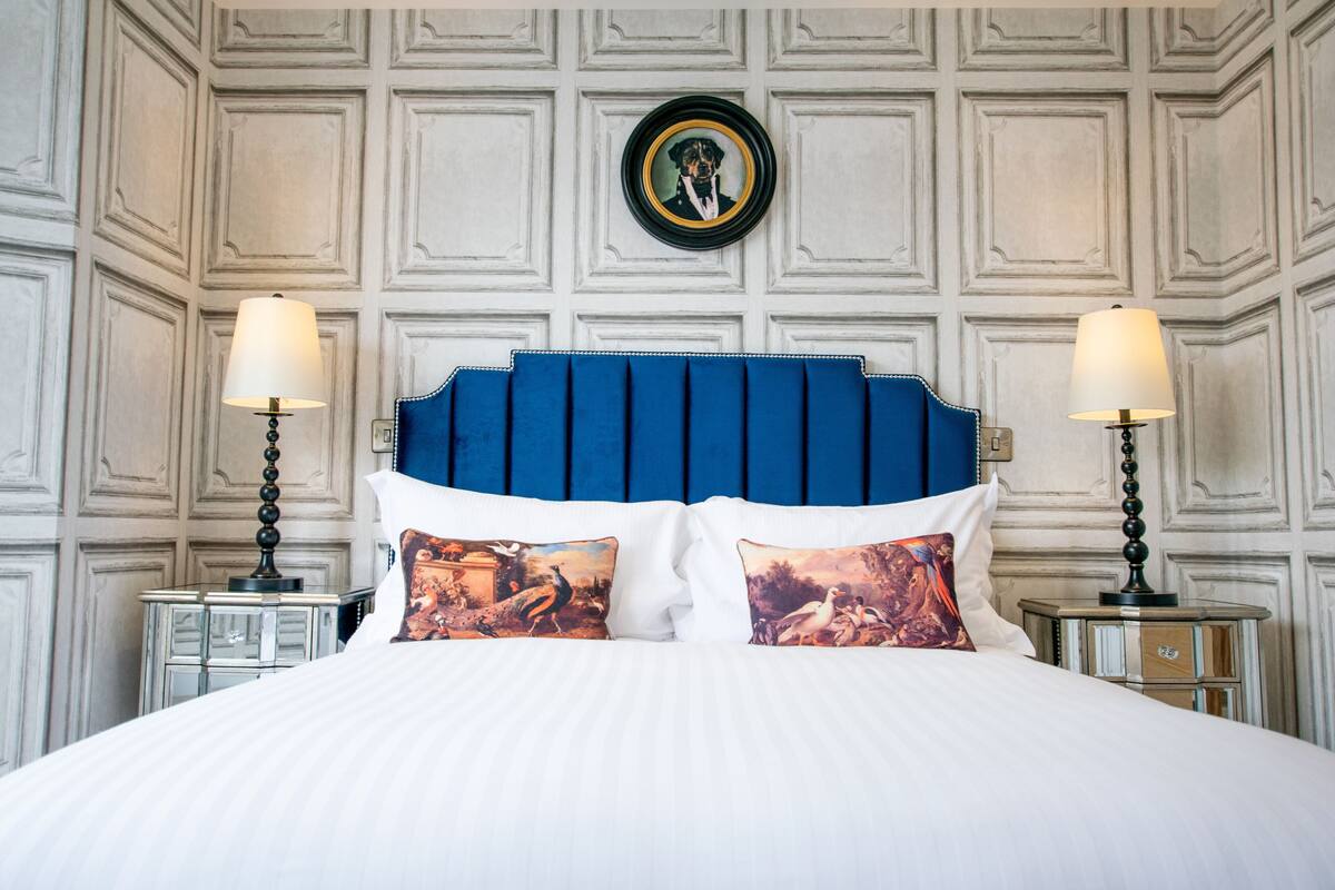 A neatly made bed with a blue headboard, two bedside tables with lamps, and a wall with a decorative panel.