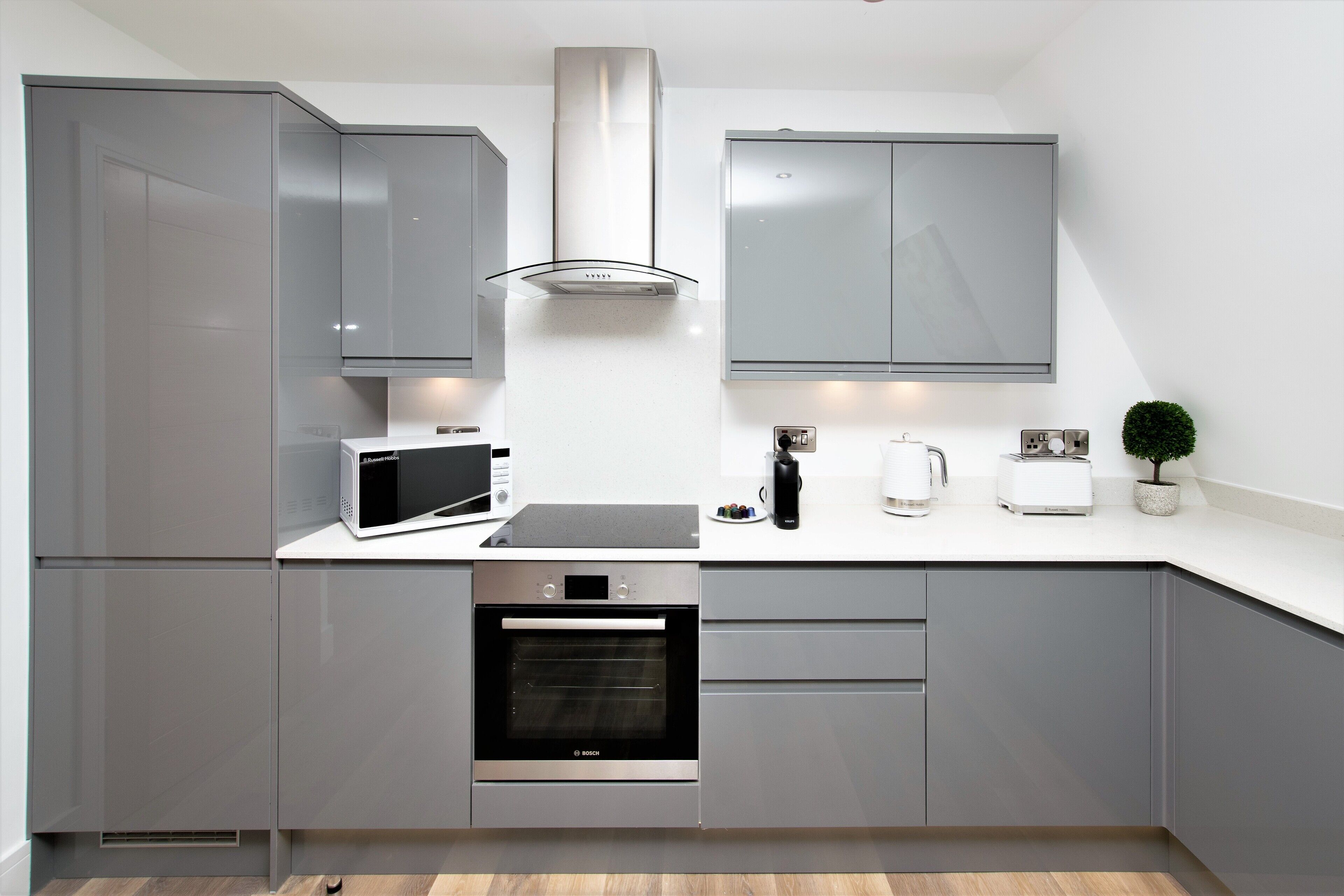 A modern kitchen with grey cabinets, a stainless steel range hood, a Bosch oven, a microwave, a coffee maker, and a small potted plant.