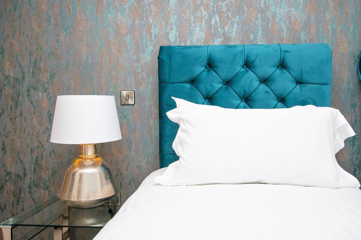 A bed with a teal headboard, a white pillow, and a metallic lamp on a bedside table against a textured wall.
