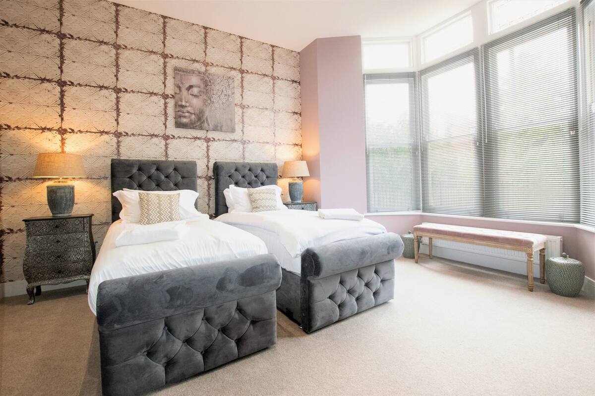 A bedroom with a large bed, two bedside tables, a bench, and a window with blinds.