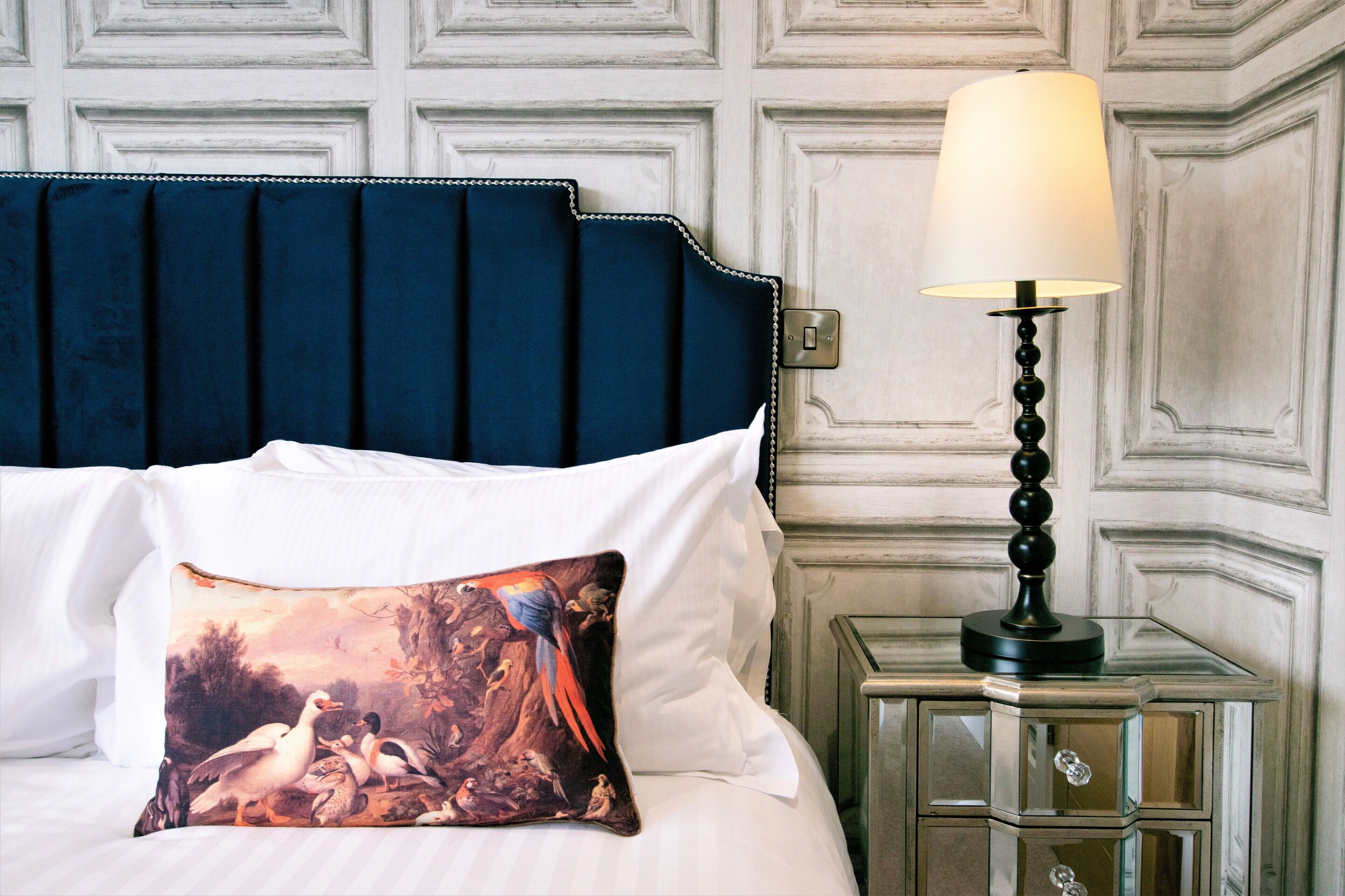 A bed with a dark blue headboard, white pillows, and a decorative pillow with a nature scene. A bedside table with a lamp is also visible.