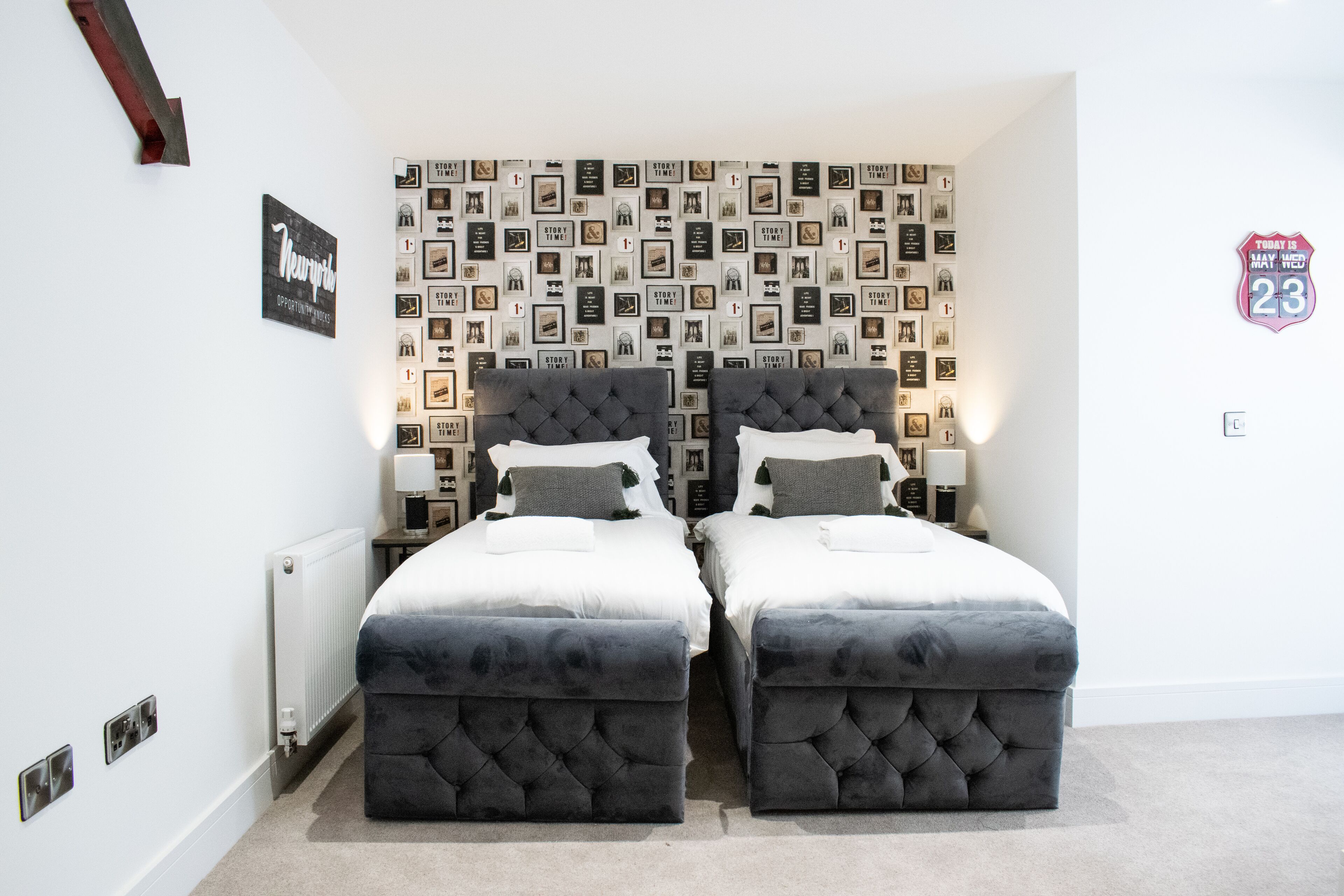 Een tweepersoonsbed met een gestoffeerd hoofdeinde en voeteinde, een wandklok en een lijst met foto's aan de muur.