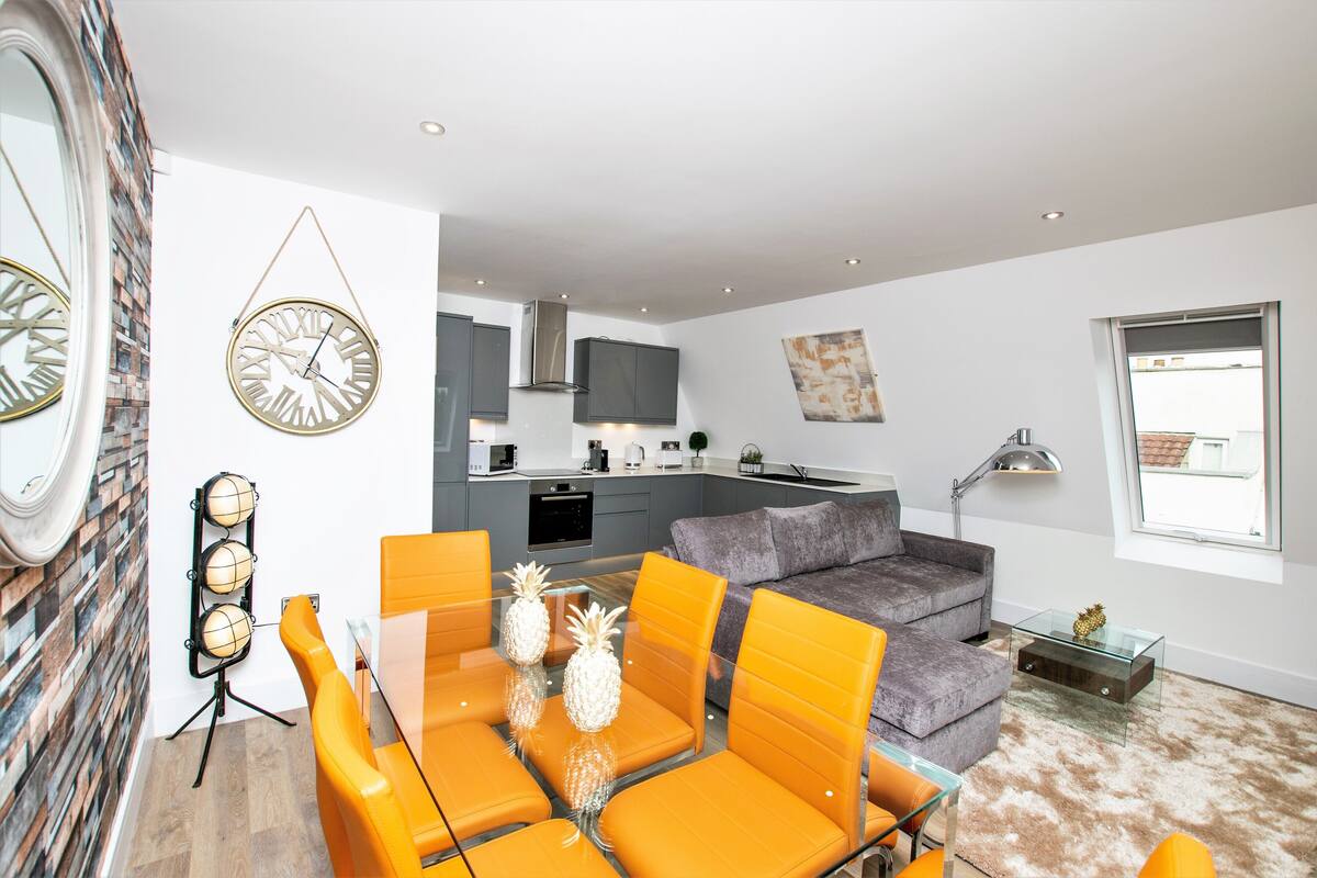 A modern living room with a glass dining table, orange chairs, a grey sofa, and a kitchen in the background.