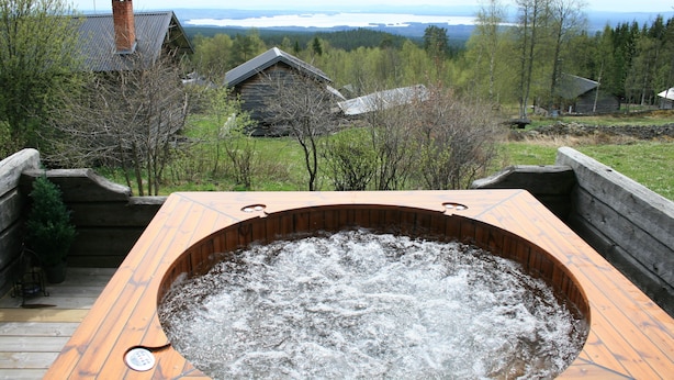 Een hottub met een houten terras, omgeven door een houten omheining, met uitzicht op een bebost gebied met huizen en een verre bergweergave.