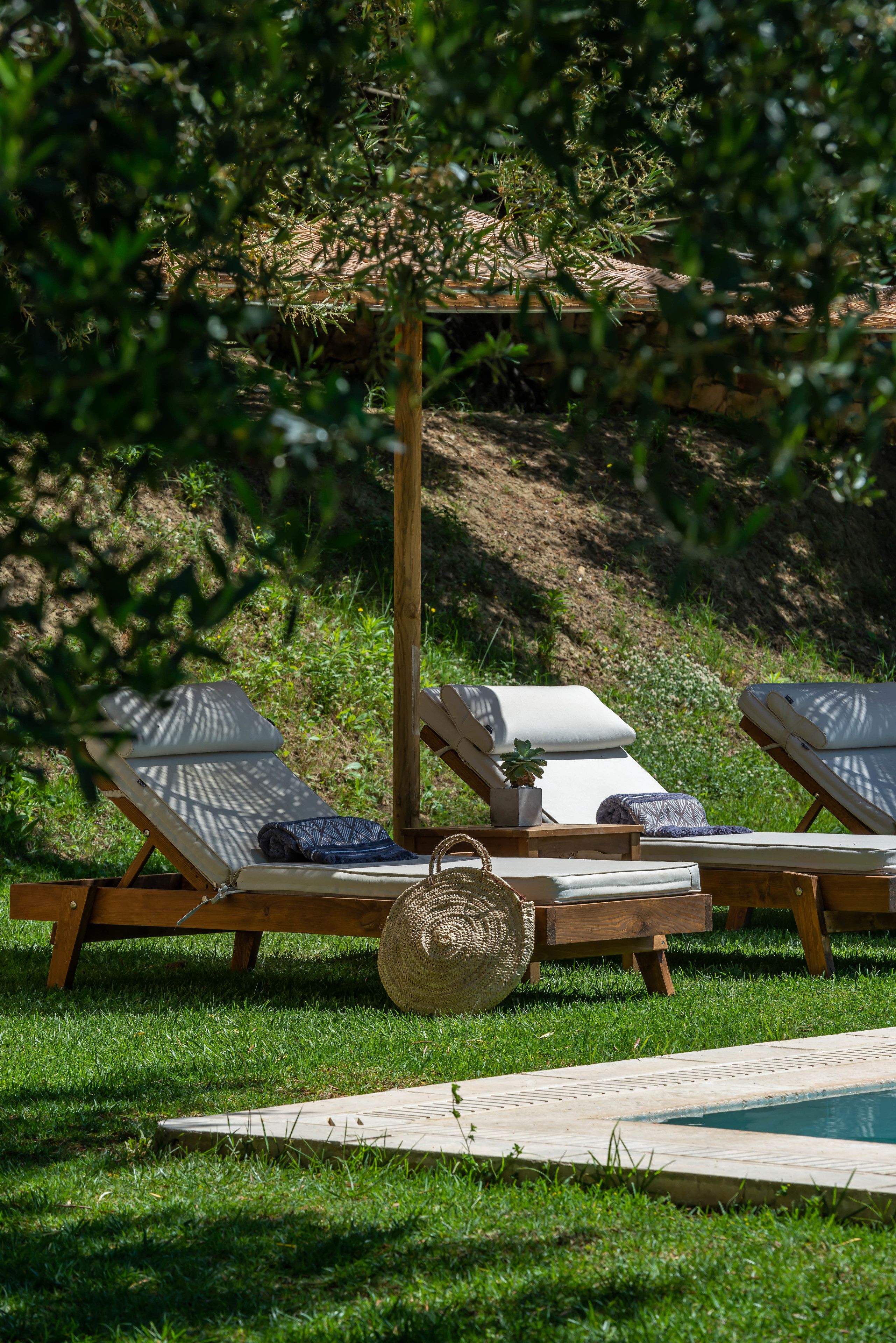 Un área junto a la piscina con tres tumbonas, una mesita con una planta y una cesta tejida.