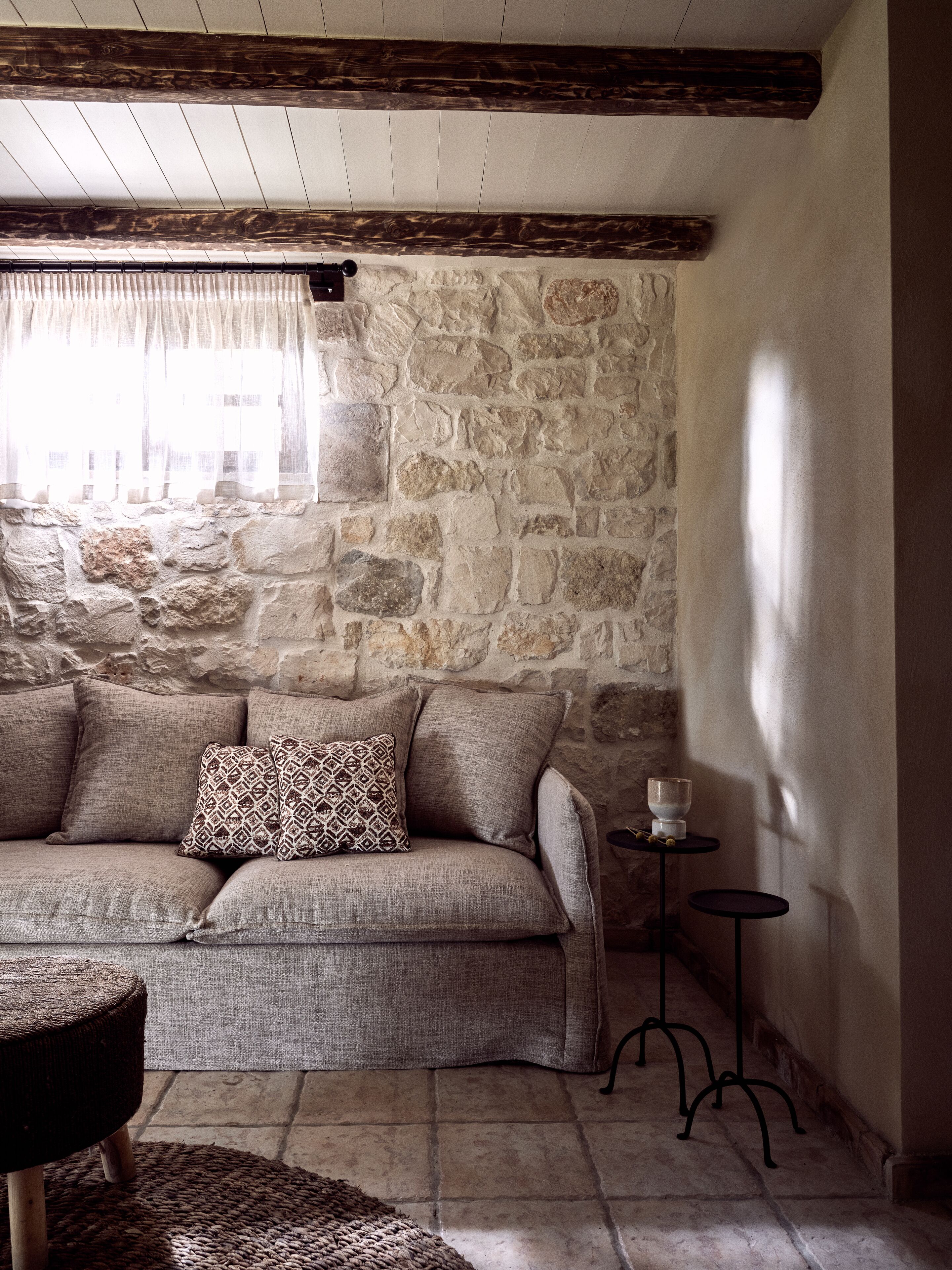 Una habitación con pared de piedra, un sofá con cojines estampados, una mesita con un vaso y una alfombra tejida.