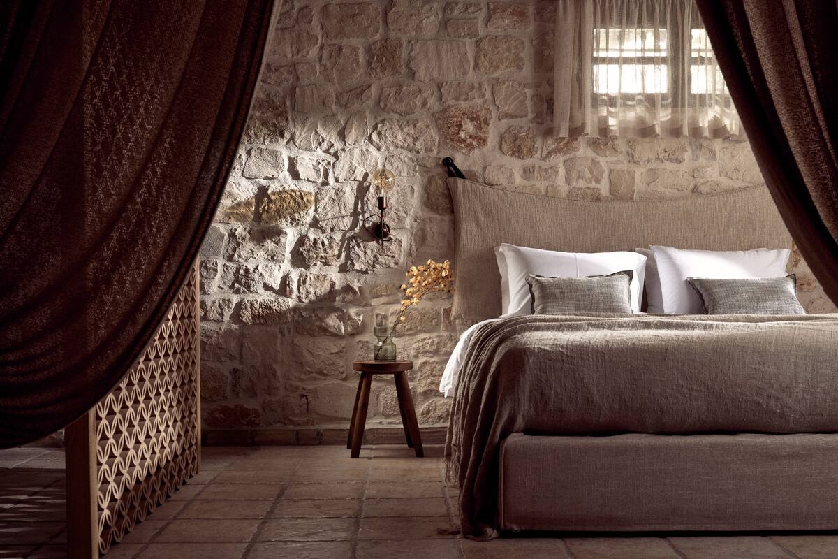 A bedroom with a stone wall, a bed with multiple pillows, a small table with a vase, and a window with curtains.