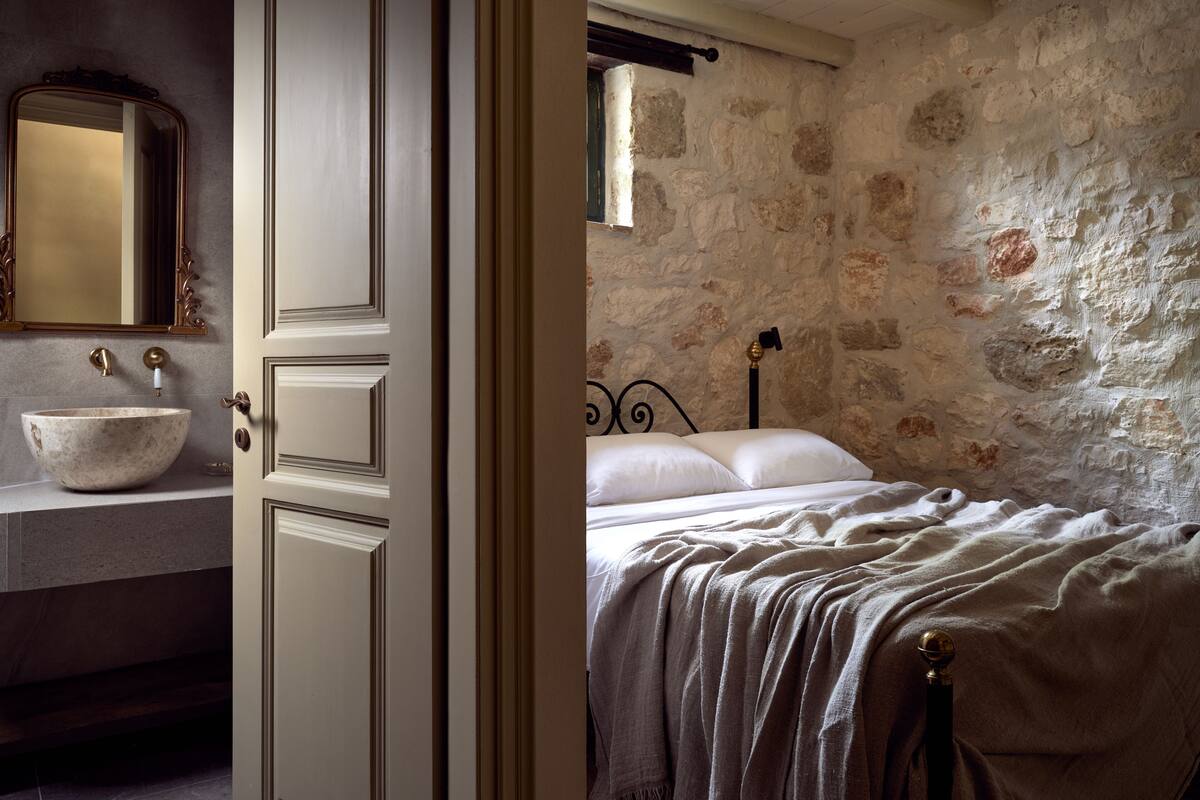 A room with a stone wall, a bed with a white bedspread, a wooden door, and a bathroom with a marble sink and a mirror.