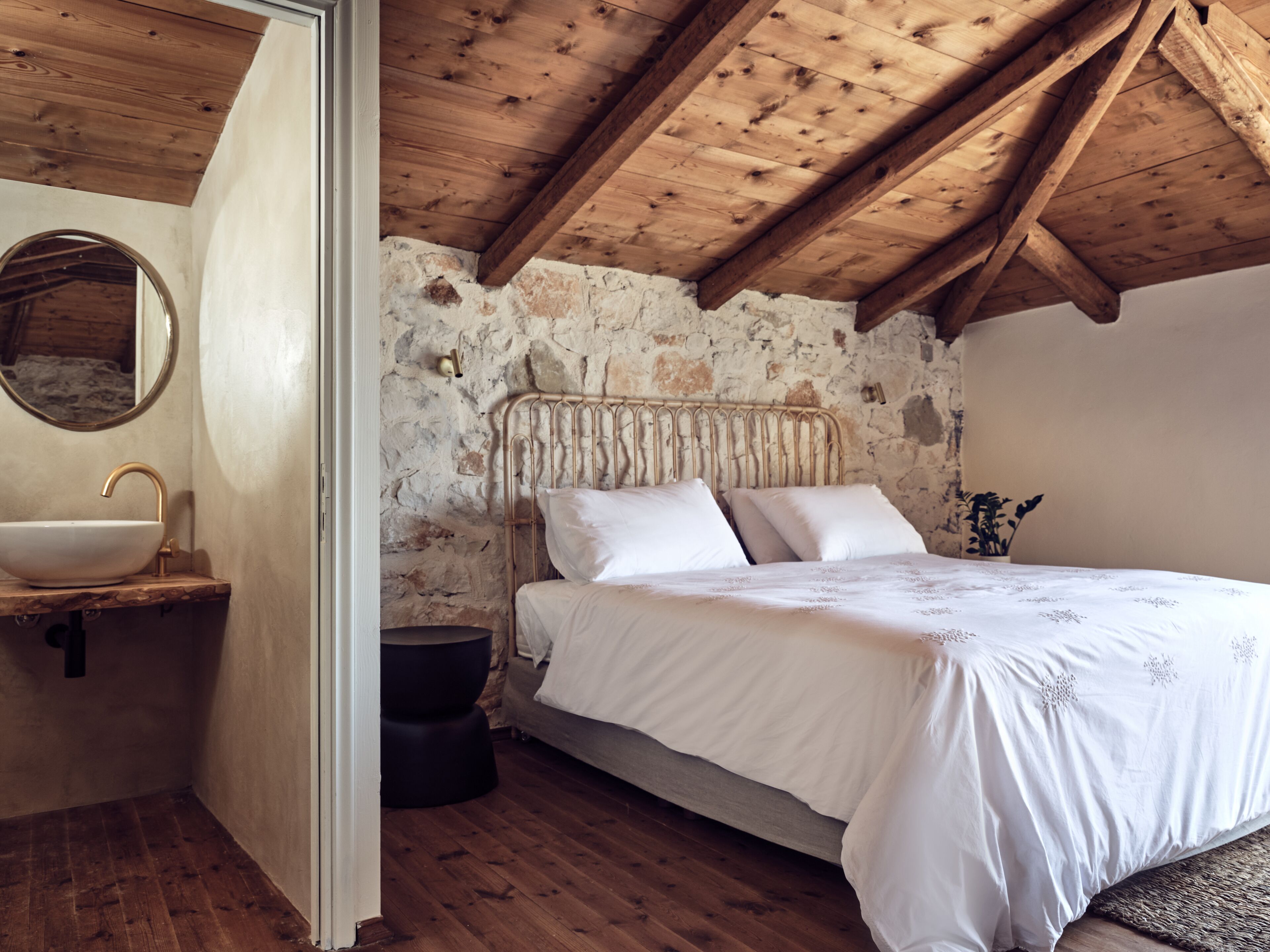 Ein Schlafzimmer mit Steinwand, Holzbalkendecke, einem Bett mit weißen Leinen, einem runden Spiegel und einem Waschbecken.